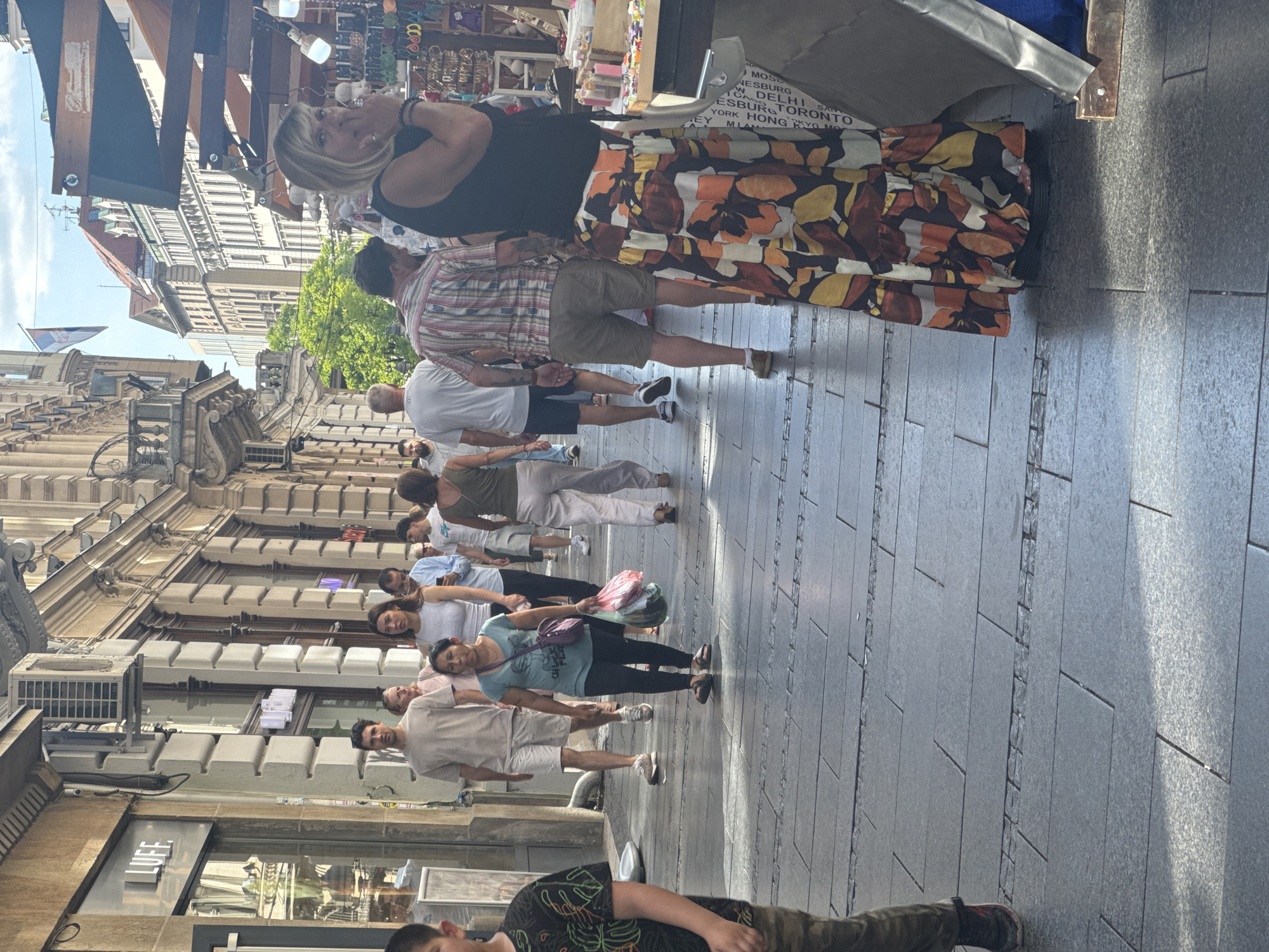 This is a vertical, medium-shot photograph of a busy pedestrian street scene in Belgrade, Serbia. Several people are walking and browsing at market stalls under an awning. In the foreground, a woman in a black tank top and a colorful, floral-patterned skirt is looking towards the camera. Behind her, a man in a plaid shirt and shorts is walking. Further down the street, a diverse group of people are visible, some in casual summer attire. The architecture of the surrounding buildings suggests a historic European city. The lighting indicates it is daytime, likely sunny with some shadows cast by the awnings and buildings. One stall has text visible, which appears to be a list of city names including "TORONTO," "HONG KONG," and "TOKYO." The overall atmosphere is lively and active, typical of a market or shopping street on a pleasant day.