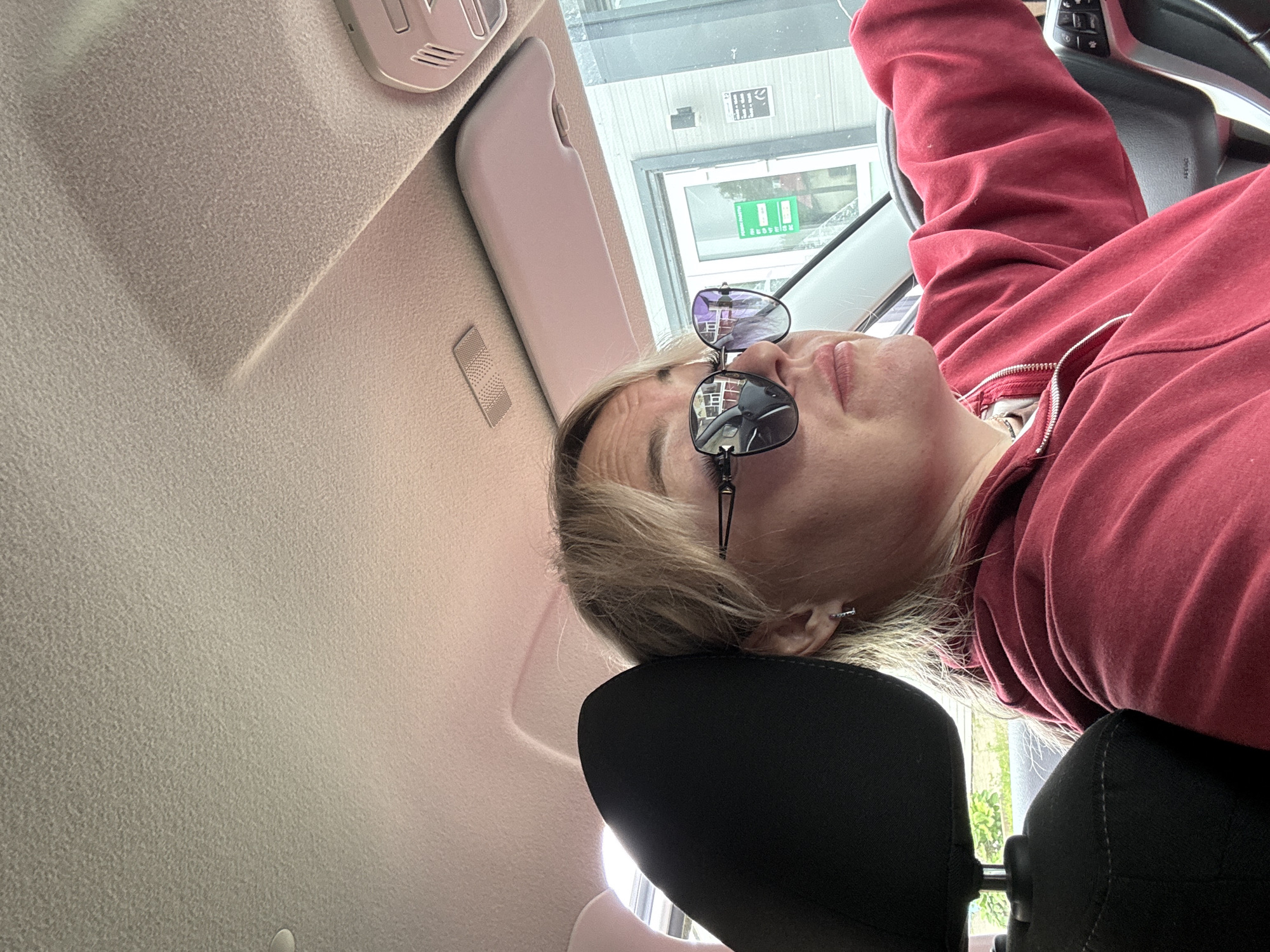In Chastyye, Russia, a blonde woman is driving a car. She is wearing sunglasses and a red hoodie. The interior of the car is visible, including the headrest of the passenger seat and the dashboard. Outside the car, a building with a sign in Russian is visible.