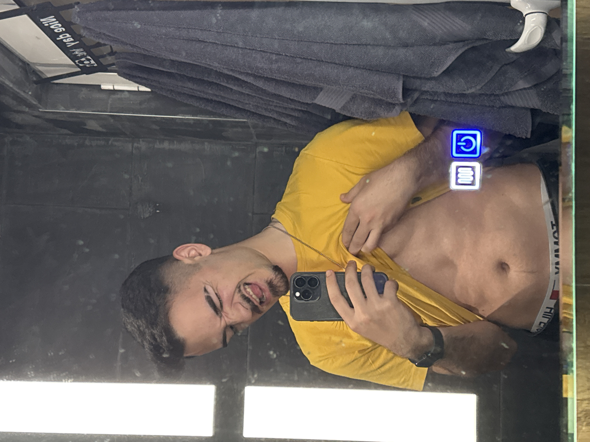 A man takes a selfie in a bathroom mirror. The man has a mustache and is wearing a yellow shirt and a gold necklace. His underwear band is visible. The bathroom has dark gray tiles, a towel rack, and an illuminated mirror with touch controls. The location cannot be determined from this image.