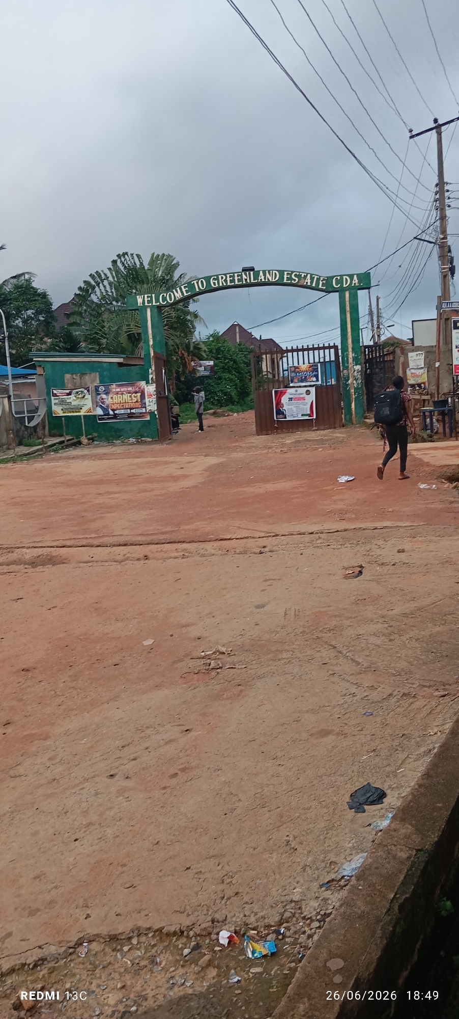 The image shows the entrance to Greenland Estate CDA in Idimu, Nigeria, on a cloudy day. A green archway at the entrance reads "WELCOME TO GREENLAND ESTATE CDA." To the left of the entrance, there is a green building with several posters on its wall, including one with a picture of a man and the text "EARNEST EXPECTATIONS." A person wearing a backpack walks away from the entrance towards the right side of the frame. Another person, seemingly a young man, stands near the entrance, facing towards the estate. The ground in front of the entrance is unpaved and appears to be dirt or gravel. There are some scattered pieces of trash on the ground and near a concrete barrier on the right side of the image. Electrical wires are visible in the upper portion of the image, connected to a utility pole. The overall scene suggests a residential area entrance with some ongoing activities or events advertised by the posters.