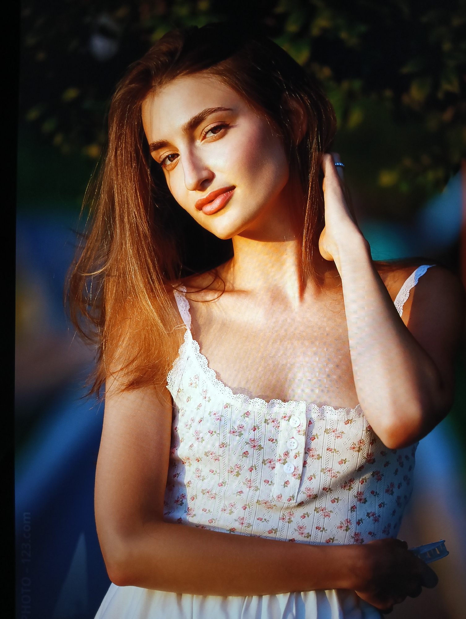 A young woman stands outdoors in sunlight, wearing a white floral patterned top with lace trim and a white skirt. She is fair-skinned with brown hair that is shoulder-length and slightly tousled. Her right hand is raised, with her fingers near her hair and a ring visible on one finger. The background features blurred foliage and what appears to be a pathway. The watermark "PHOTO-123.com" is at the bottom left corner of the image. There is no evidence in the image to support the assertion that the location is in the city of Nha Trang Ward, Vietnam.