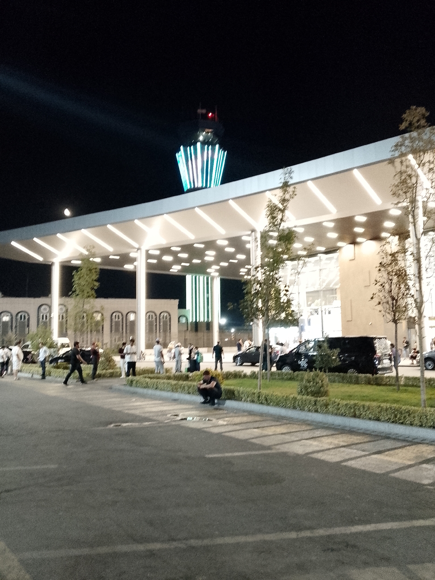 The image depicts a night scene at the Tashkent International Airport in Tashkent, Uzbekistan. A modern, illuminated terminal building with a white canopy and strip lighting is visible. In the background, a distinctive tower, likely an air traffic control tower, is lit with green and white vertical stripes. Several people are present in the foreground and midground, some walking and others standing. A person is crouched down in the lower right of the frame. Several vehicles, including a black van and a car, are parked in the area. The ground is paved, and a grassy area with hedges and trees lines the edge of the paved area. The overall impression is one of an active airport environment at night.