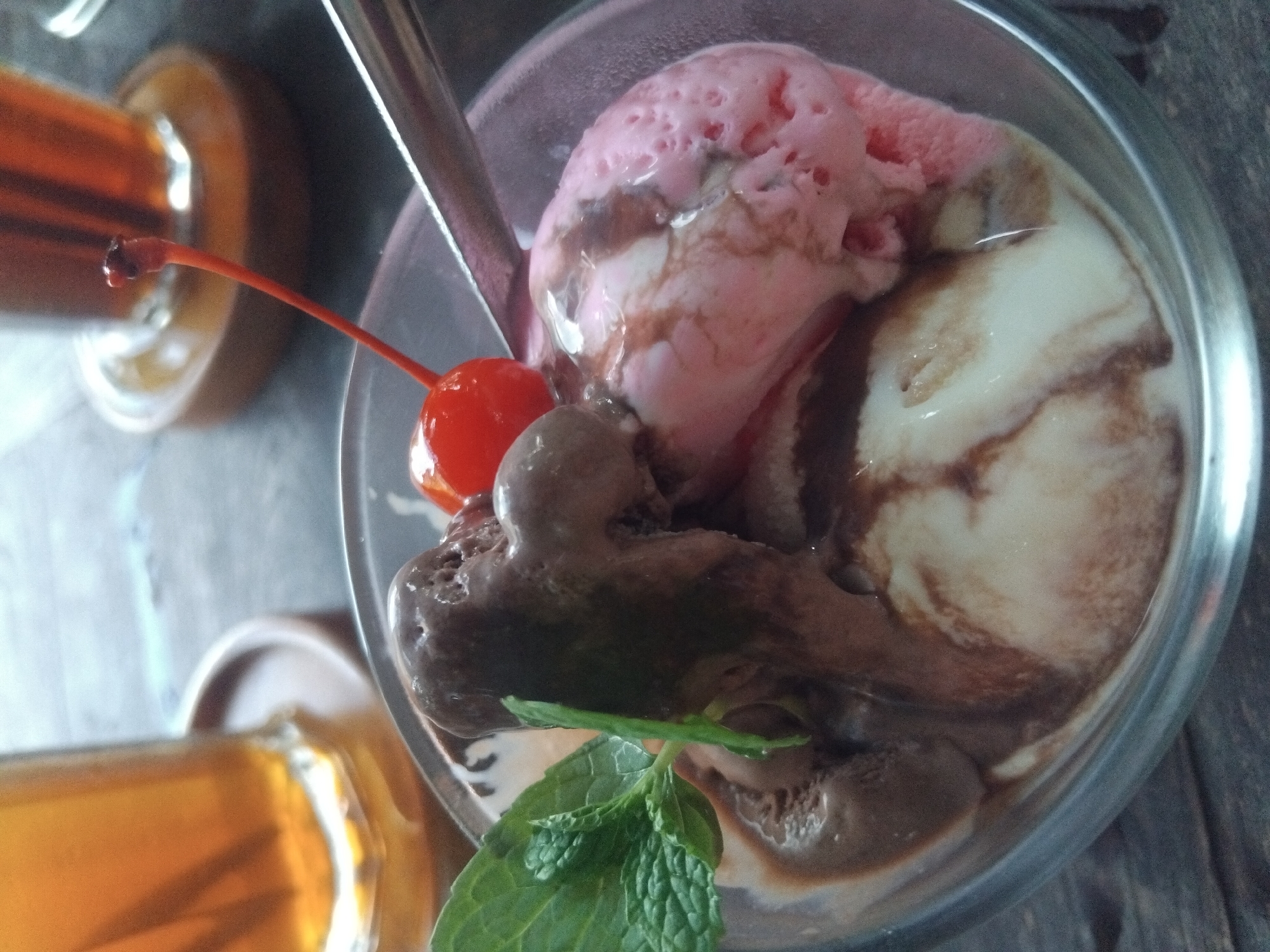 A close-up shot shows a glass containing a dessert with three scoops of ice cream: strawberry, vanilla with chocolate swirl, and chocolate. A bright red cherry with a long stem sits atop the ice cream, next to a stainless steel spoon. A sprig of green mint leaves garnishes the dessert. Two glasses filled with a translucent, amber liquid (likely tea or juice) are partially visible in the upper left and lower left of the frame. The scene appears to be set indoors on a wooden surface. The location is Unknown, Unknown.
