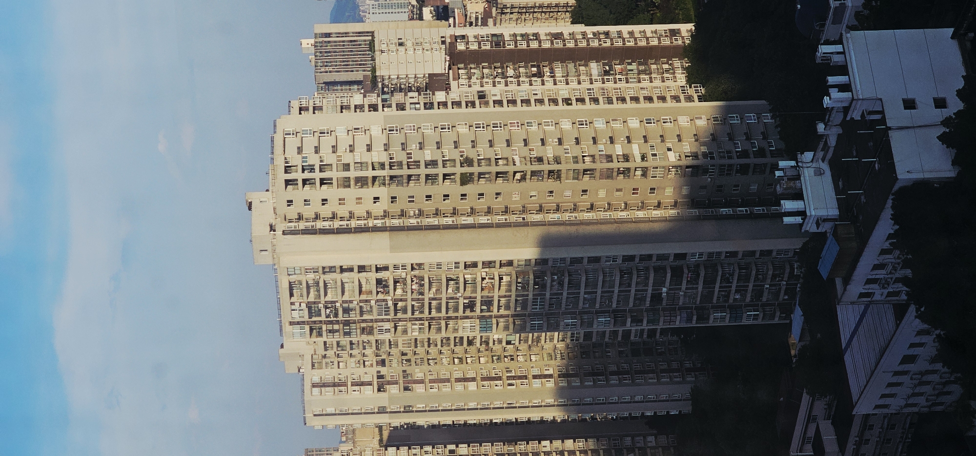 A wide shot, taken from an elevated perspective, captures a dense urban cityscape in Jiulongpo District, China. The primary subject is a towering, cream-colored apartment building that dominates the center of the frame, its facade marked by numerous windows and balconies. A distinct shadow stretches vertically down the left side of this building, indicating the direction of the sun. To the right of this main structure, other multi-story buildings of varying heights and architectural styles are visible, receding into the background. Some of these buildings also feature visible windows and balconies, suggesting a high population density. In the foreground and scattered amongst the buildings are dark green trees, their foliage providing a contrast to the urban architecture. The sky above is a clear, light blue, with faint white clouds scattered across it. The overall impression is of a sunlit urban environment, possibly in the late afternoon due to the long shadow.