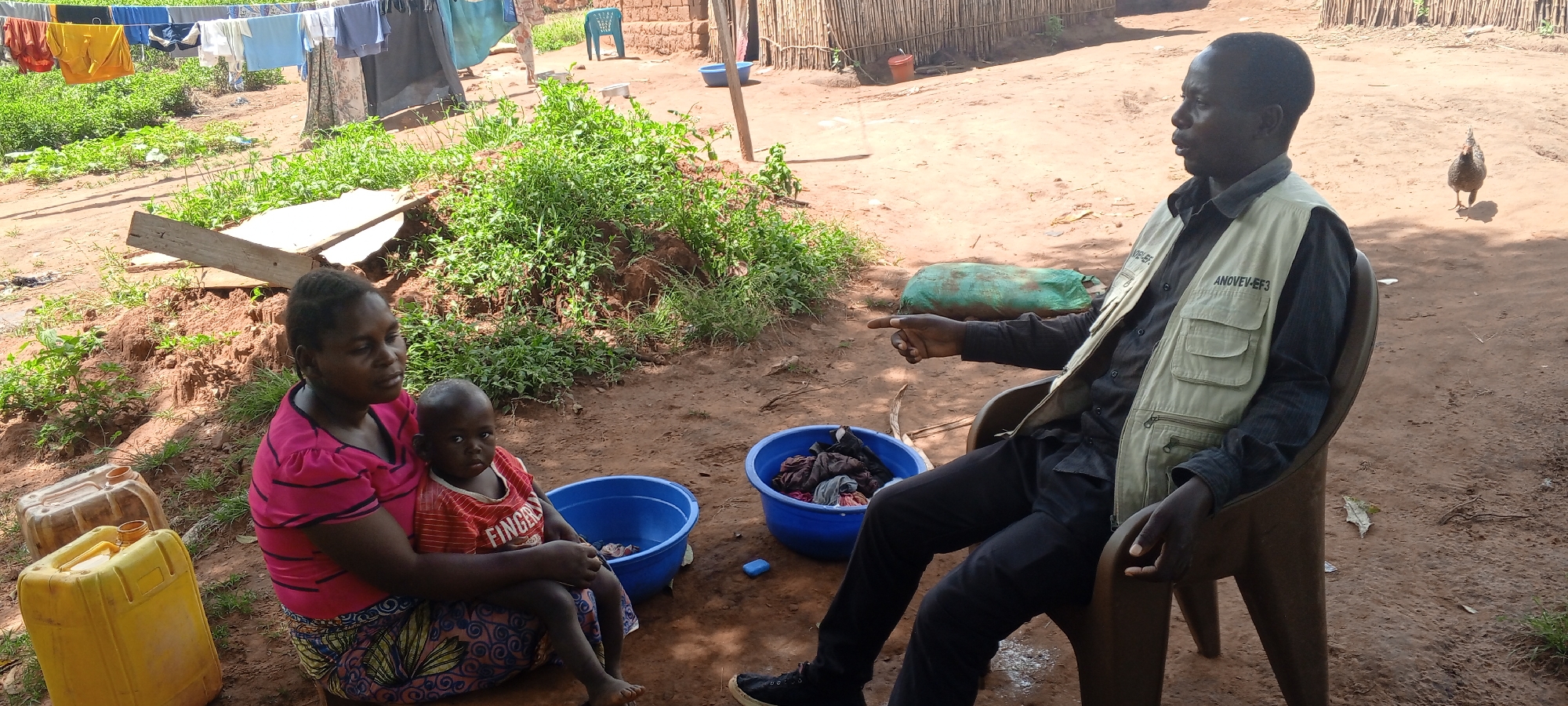The image shows a man sitting on a chair in an outdoor setting in Makungu, Democratic Republic of the Congo, apparently engaged in a conversation with a woman who is sitting on the ground, holding a young child. The man is wearing a black shirt, black pants, and a light-colored vest with the text "ANOVEV-EF3" on it. He is gesturing with his right hand. The woman is wearing a pink shirt with red sleeves, a patterned skirt, and is holding a child in a red shirt that says "FINGER". Two blue plastic tubs are on the ground near the woman, one empty and one filled with clothes. Beside the woman are three yellow plastic jerrycans. The background includes a clothesline with laundry hanging, a wooden fence, and a chicken. The ground appears to be dirt.