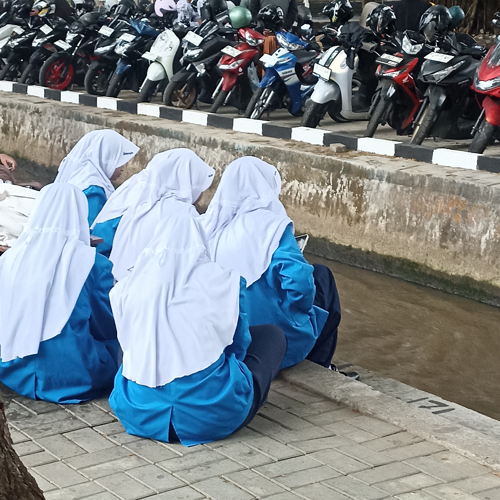 The image depicts a row of parked motorcycles behind a low concrete barrier. In front of the barrier, four individuals wearing blue uniforms and white headscarves are sitting on a paved walkway, facing away from the camera. The individuals appear to be young women, possibly students, and at least one of them is looking at a mobile phone. The background consists of more parked motorcycles and a dense arrangement of trees and foliage, suggesting an outdoor environment. The scene is likely located in a public area such as a parking lot or a school vicinity in the city of Magelang, Indonesia. The motorcycles are predominantly modern models in various colors including red, blue, black, and white. The concrete barrier has a black and white striped pattern along its top edge. The water visible in the lower right portion of the image indicates a canal or ditch.
