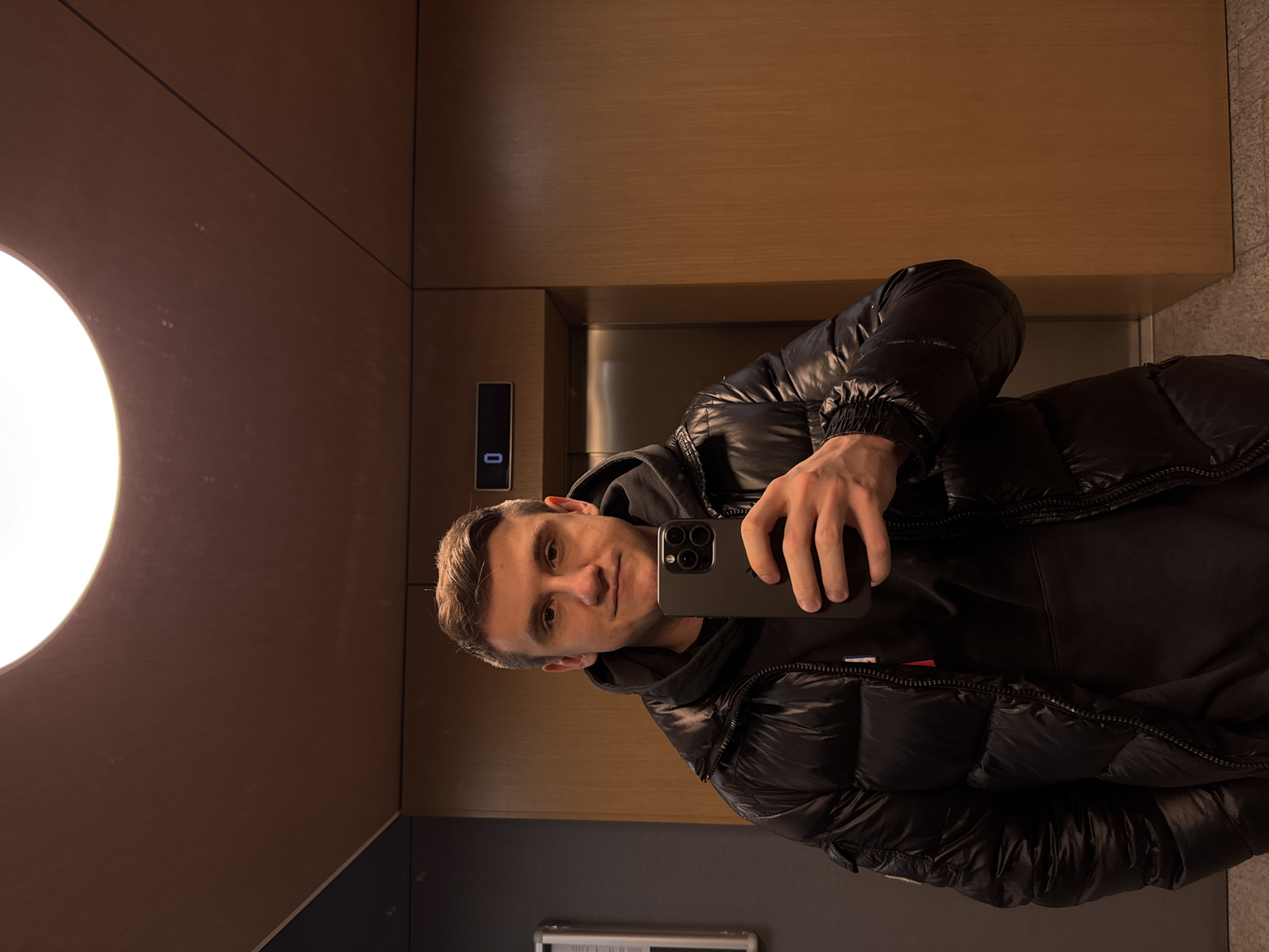 A man is taking a selfie inside an elevator. He is wearing a black puffy jacket over a black hoodie. The elevator has wooden paneling and a round light fixture. The elevator button panel reads "0". A small label with red, white, and blue stripes is visible on his jacket. The location is likely Warsaw, Poland, due to the man's attire and the modern elevator design being common in urban environments.