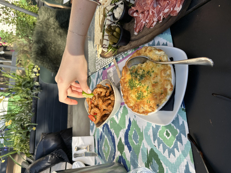 The scene is an outdoor dining area within the city of London, United Kingdom. A dark table is partially covered by a patterned textile featuring geometric designs in shades of blue, green, and white. A human hand, appearing to be female, is positioned centrally, actively squeezing a lime wedge over a white bowl filled with fried calamari, garnished with red pepper slices. To the lower center, a white oval gratin dish contains a baked, cheese-topped entree, garnished with green herbs, with a silver spoon resting within it. To the right, a wooden serving board displays an array of sliced cured meats, rectangular cheese slices drizzled with balsamic glaze, flatbread segments, and two small black containers holding gherkins and pearl onions. Further to the left, a silver jug and a small white block with a knife and fork are visible. The background consists of dark planters containing various green plants and yellow flowers, suggesting a patio or terrace environment under natural daylight.