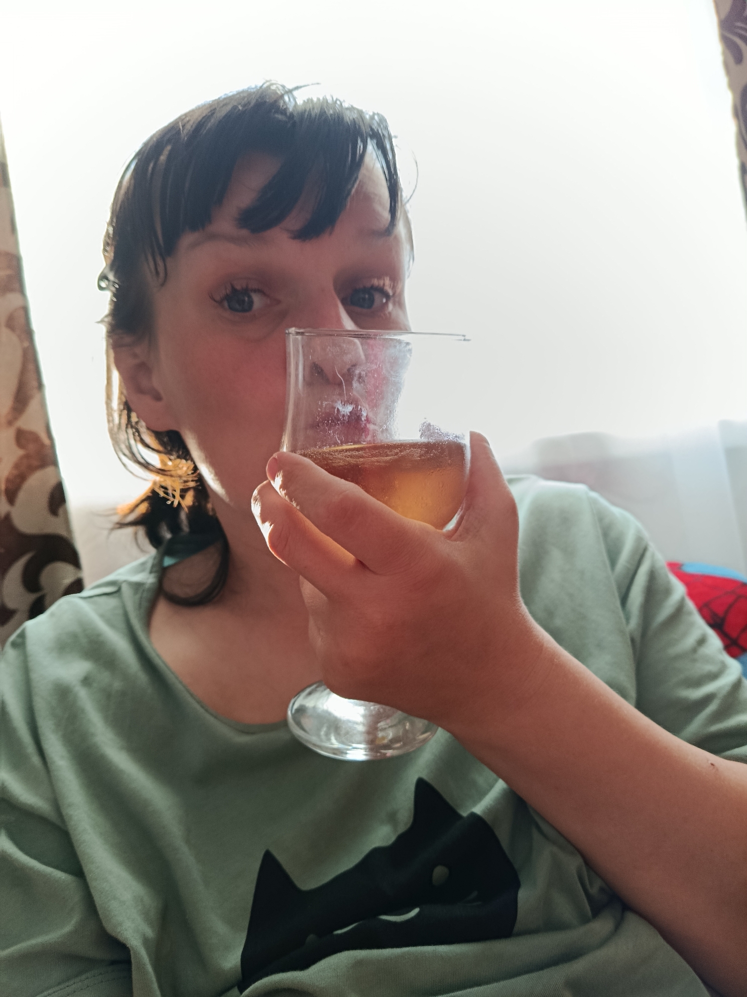 A person with dark hair and bangs is holding a wine glass with a light-colored liquid, possibly beer or wine. The person is wearing a green t-shirt with a black cat design. The background is bright, with a curtain visible on the left and a window on the right. The scene appears to be indoors. There is no indication that the image was taken in the city of Фоки, Russia.