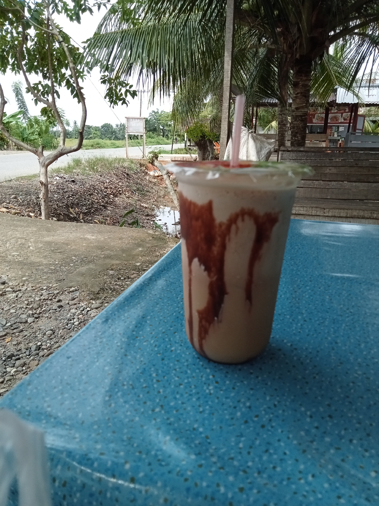 A plastic cup filled with a light-colored beverage, possibly a milkshake or smoothie, with streaks of chocolate syrup drizzled down the sides. The cup has a clear plastic lid with a pink straw inserted. The cup is placed on a blue speckled table in the foreground. In the background, there is a wooden structure, possibly a bench or seating area, with some posters or signs attached. Palm trees and other foliage are visible, suggesting a tropical or subtropical environment. The ground appears to be a mix of gravel and dirt. A road is visible in the distance. The scene is likely outdoors, possibly at a roadside stall or cafe in Matangsijuek Timur, Indonesia. There are no people clearly visible in the image.