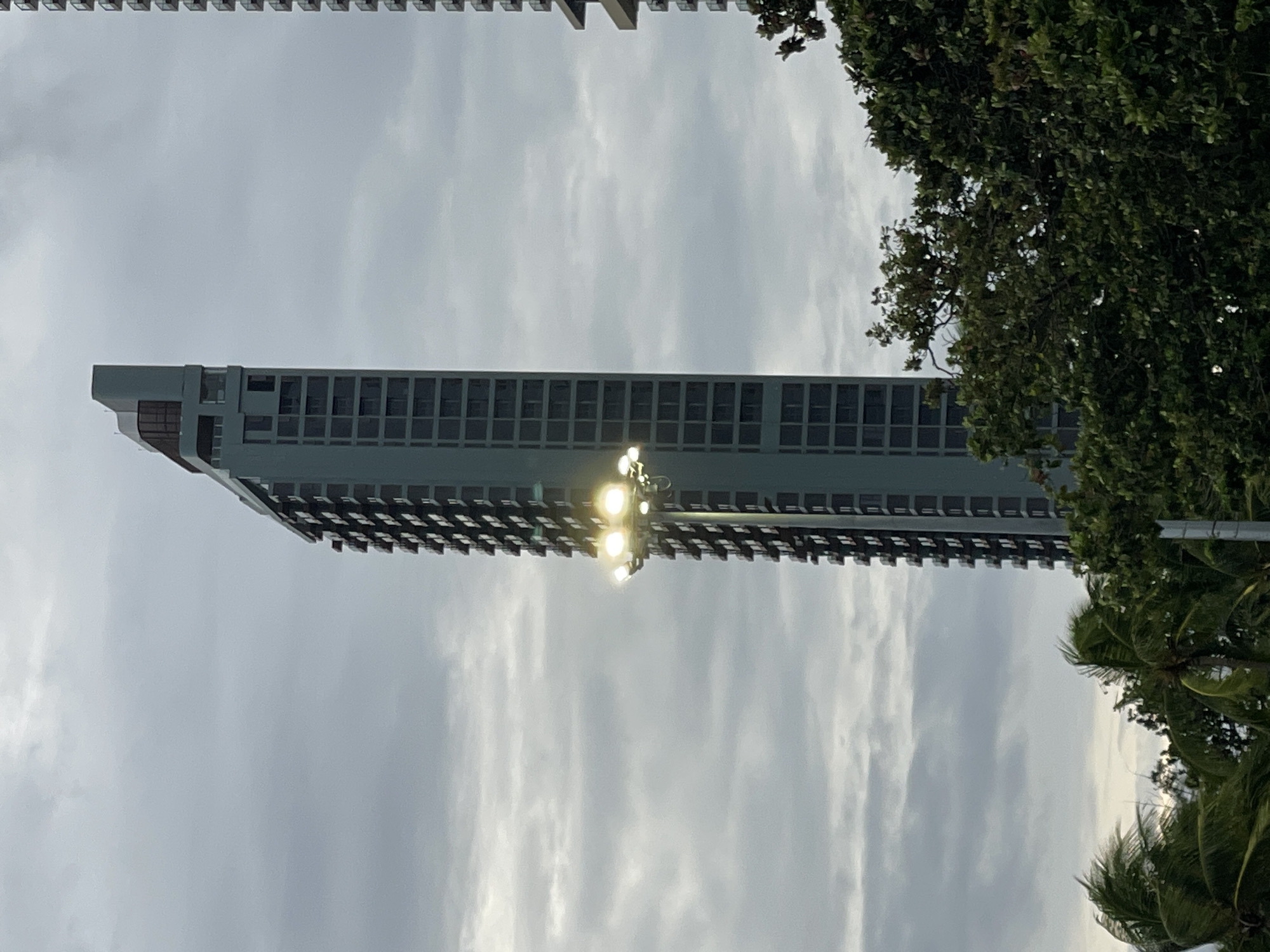 The image shows a tall, modern condominium building with a light blue facade and numerous balconies. The building is partially obscured by lush green trees in the foreground. A cluster of bright spotlights is attached to a pole extending from the side of the building, illuminating the cloudy sky. In the background, another similar condominium building is visible. The scene appears to be in a tropical or subtropical urban environment, likely in Pattaya City, Thailand, given the architecture and vegetation. The lighting suggests it might be dusk or an overcast day. There are no people visible in the image.