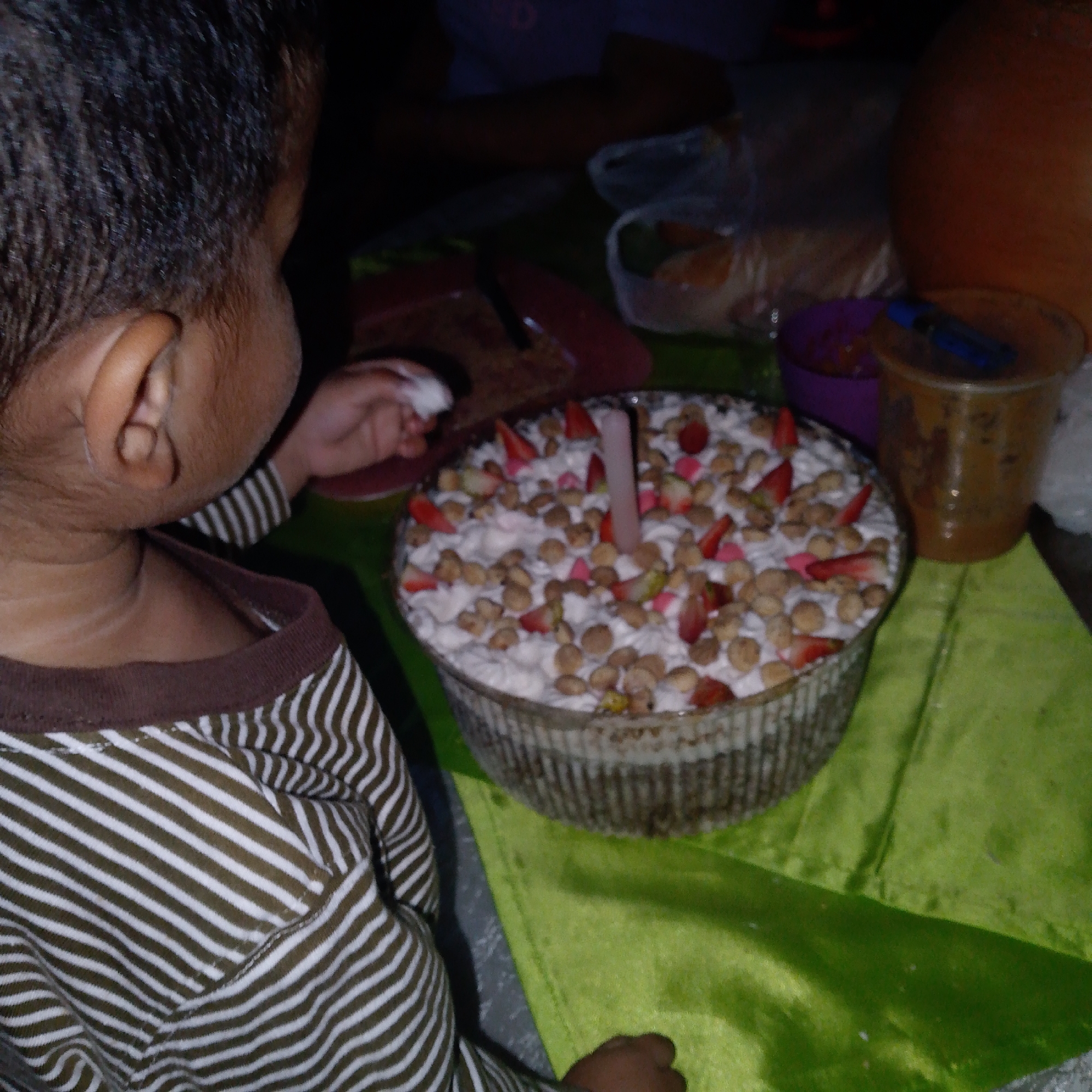 The image captures an intimate, dimly lit scene, likely indoors during the evening or night. The primary subject is a young child, seen from behind and to the side, with dark, short hair and visible ear. The child is wearing a brown and white striped long-sleeved shirt and appears to be holding a small piece of white material (possibly cotton or tissue) in their right hand.
In the foreground, a large, glass bowl holds a dessert or cake with a white, creamy topping, adorned with sliced strawberries, small pink heart-shaped candies, and numerous light brown, round cereal or cookie pieces. A single, tall, slender pink candle is prominently placed in the center of the dessert, suggesting a celebration like a birthday.
The dessert bowl rests on a vibrant lime-green tablecloth. Surrounding the dessert are several other items: a partially visible red plate, a small purple bowl, and a clear plastic cup containing a dark beverage with a blue pen or utensil sticking out. In the background, other items like plastic bags holding food and a large, round, reddish-brown pot or container are dimly visible, further indicating a gathering or mealtime setting. The overall atmosphere suggests a casual, perhaps celebratory, event. There is no visible text in the image. The location context provided is Tinaquillo, Venezuela.