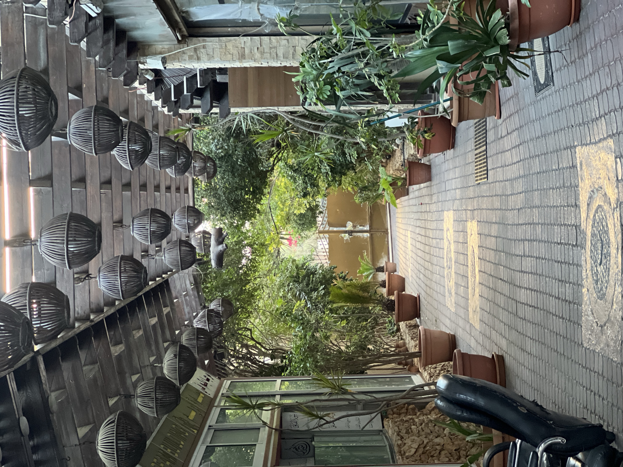 The image depicts an outdoor pathway, likely a garden or patio area, in the city of Gaza, Palestinian Territories. The pathway is paved with light-colored bricks arranged in a basketweave pattern, interspersed with yellow rectangular markings and circular metal grates.
Hanging from a wooden overhead structure, which appears to be a pergola or covered walkway, are numerous spherical, woven lamps. These lamps are dark in color and vary in size. The overhead structure itself is made of wooden beams and planks.
Flanking the pathway are several potted plants of varying sizes and types, including tall, thin-leaved plants and broader-leafed foliage. Some plants are placed in terra cotta pots, while others are in larger, darker brown pots. The area is lush with greenery, suggesting a well-maintained outdoor space.
To the left, a glass-fronted building or structure is visible, with stone cladding at its base. On the glass, there is Arabic text printed. A portion of a black, leather armchair is visible in the foreground on the left.
In the distance, at the end of the pathway, a large, double-leafed metal gate is visible. The overall scene suggests a tranquil and shaded outdoor area, possibly a cafe, restaurant, or private residence. The lighting indicates it is daytime, with diffused sunlight filtering through the overhead structure and foliage. No people are visible in the image.