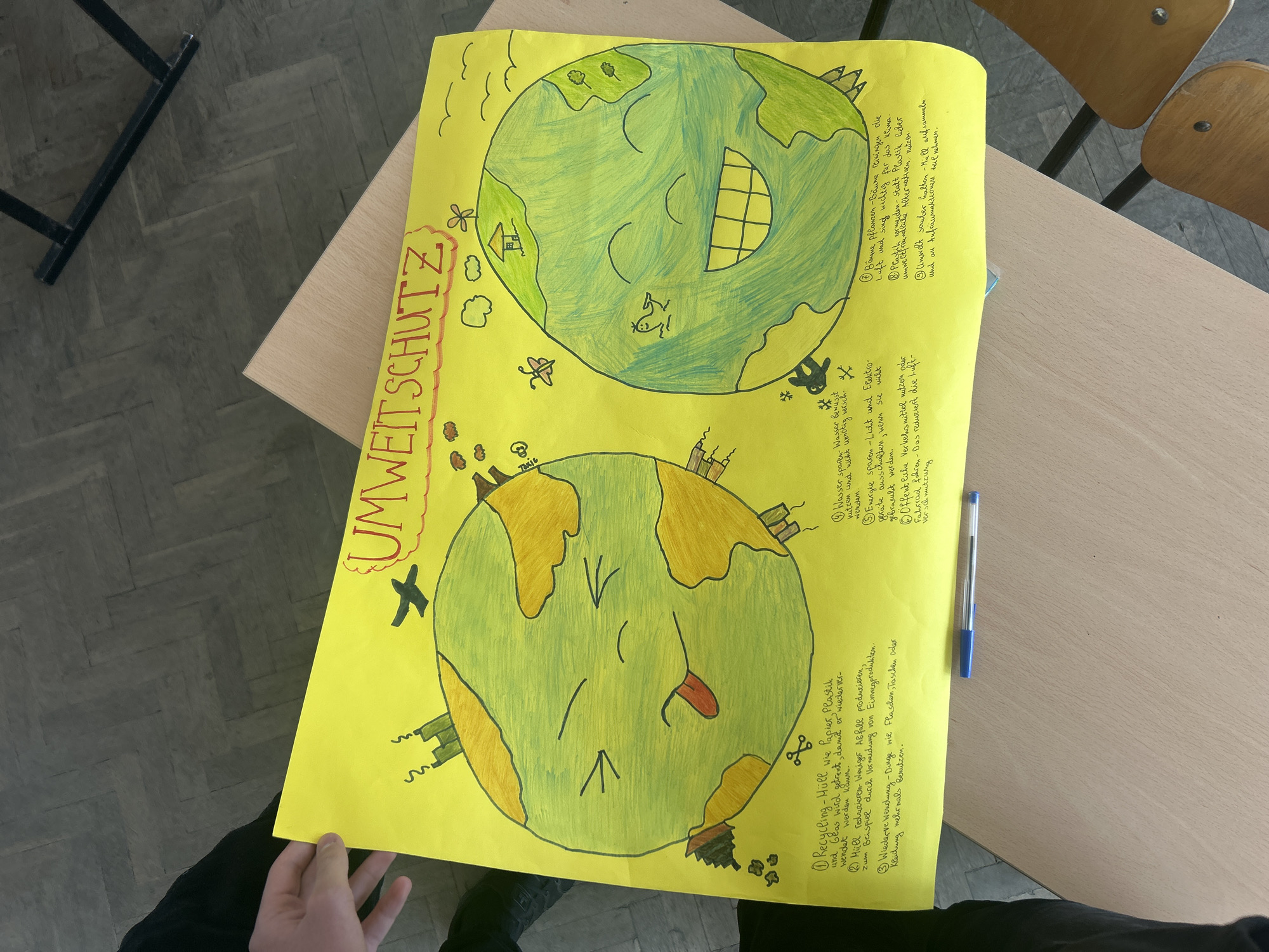 The image shows a yellow poster titled "UMWEITSCHUTZ" (environmental protection in German), placed on a wooden desk in what appears to be a classroom setting. The poster contains two drawings of the Earth, one depicted as happy and the other as sad. The happy Earth is drawn with green landmasses and has a large smile. The sad Earth is drawn with yellow landmasses, has a frowning expression, and includes depictions of pollution from factories and an airplane. Handwritten notes in German are placed around the drawings, listing ways to protect the environment, such as recycling, reducing waste, saving water and energy, and planting trees. A blue pen is also lying on the desk next to the poster. A hand is visible at the bottom left corner, seemingly holding or presenting the poster. Part of a wooden chair is visible in the upper right corner, and a desk leg can be seen on the left. The flooring appears to be a herringbone pattern.