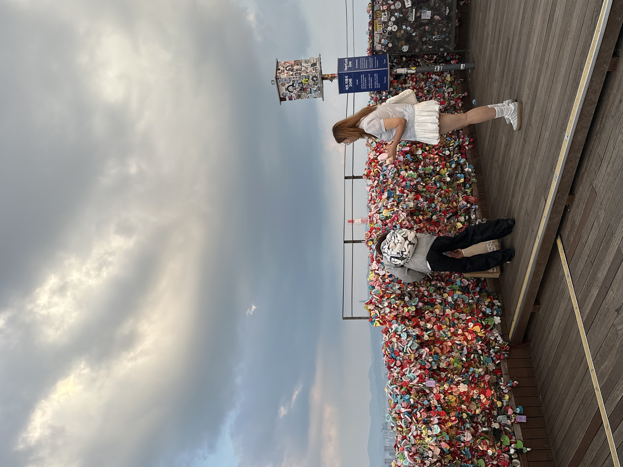 The image depicts the Namsan Seoul Tower, a landmark in Seoul, South Korea, famous for its "Locks of Love" installation. In the foreground, a wooden deck is visible, leading to a large wall covered in colorful heart-shaped locks. Two individuals are present. On the left, a person wearing a gray hoodie and black pants is bending down, seemingly placing or retrieving something from behind the locks. On the right, a young woman with long brown hair, wearing a light blue short-sleeved shirt and a white ruffled skirt, stands facing the locks, holding two pink items in her hands. A signpost with Korean text and "HeartLock Zone" in English is situated behind her. The background shows a hazy cityscape with mountains and a cloudy sky. The overall scene captures a moment at a popular tourist attraction, characterized by the symbolic display of love locks.