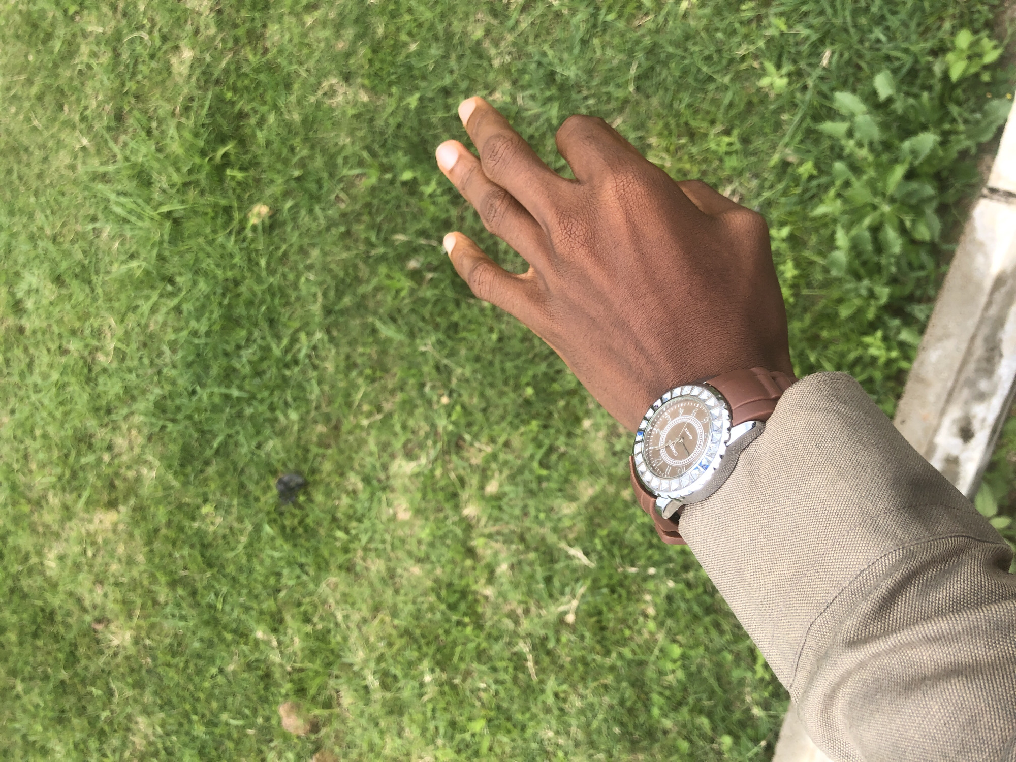 The media file shows a close-up, high-angle shot of a person's right hand and forearm, which is resting on a beige-colored fabric, likely a jacket or shirt. The hand is open, with fingers slightly splayed, and the palm is facing upwards. The person's skin tone is dark.
On the wrist, a wristwatch with a brown leather strap is clearly visible. The watch face is round and has a brown dial. The bezel of the watch is decorated with numerous small, shiny stones, possibly diamonds or crystals, and also features a rotating outer ring with markings. The brand name "CHANEL" and "QUARTZ" are visible on the watch face.
The background consists of a vibrant green grassy area with some small plants interspersed. The grass appears somewhat uneven and possibly dry in places. To the right of the arm, there is a vertical element that appears to be a light-colored concrete or stone structure, possibly a curb or a wall.
The lighting suggests it might be daytime, with ample natural light illuminating the scene. The overall impression is casual, perhaps someone outdoors in a grassy area. There are no strong emotional cues conveyed, and the activity appears to be a static pose, with the hand resting. The setting, Gwafan, Nigeria, suggests a potentially warm climate. The presence of the watch indicates a focus on timekeeping or fashion.