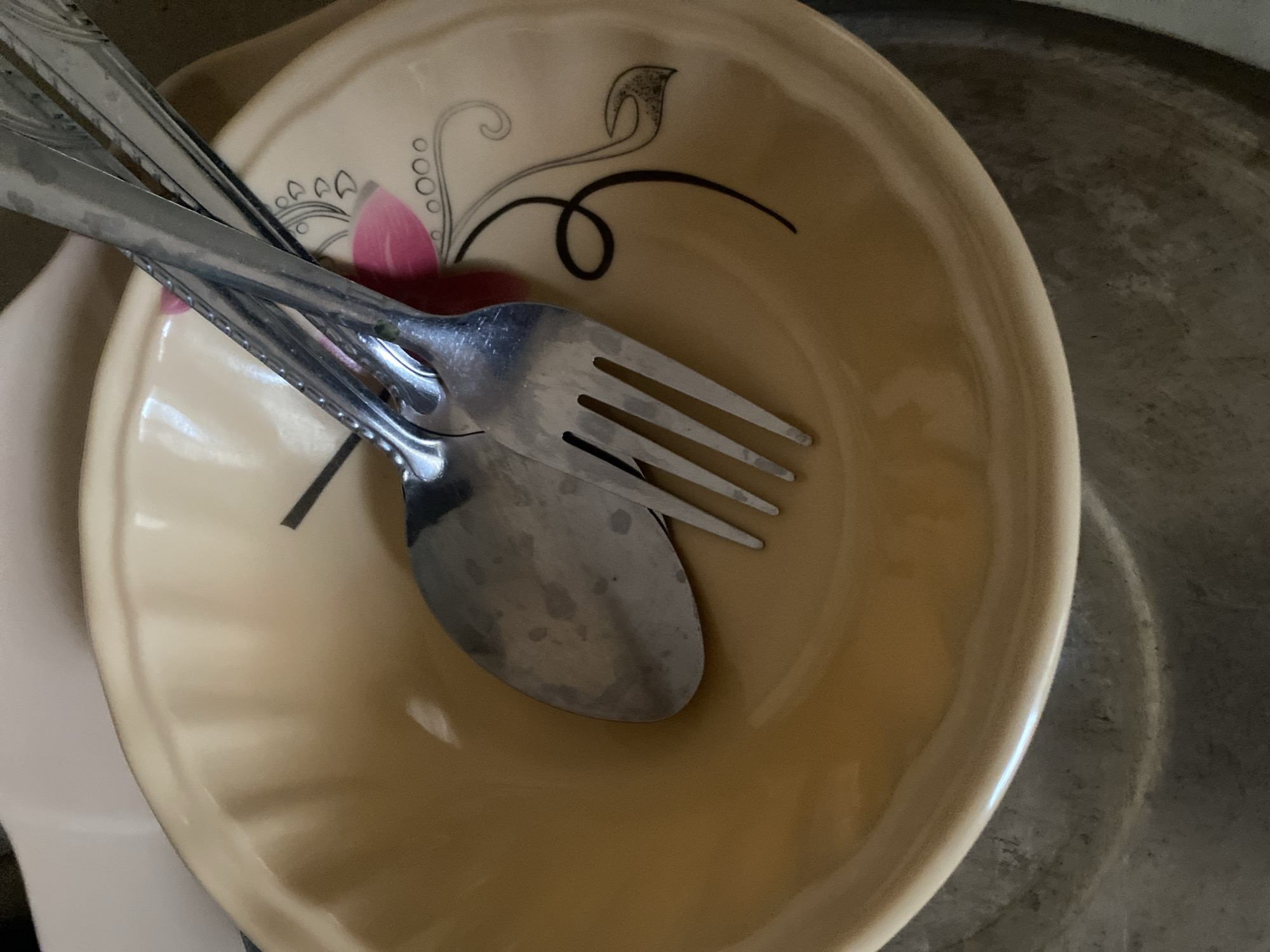 The image shows a close-up view of a light yellow bowl with a floral design, containing a spoon and a fork. The spoon is on top of the fork, and both utensils are made of silver metal. The bowl has a subtle embossed pattern and a decorative border with black and pink floral motifs. The setting appears to be indoors, with soft lighting. The image does not contain any information or context that would allow for the determination of its location within the city of Yola, Nigeria, or any other specific geographical area.