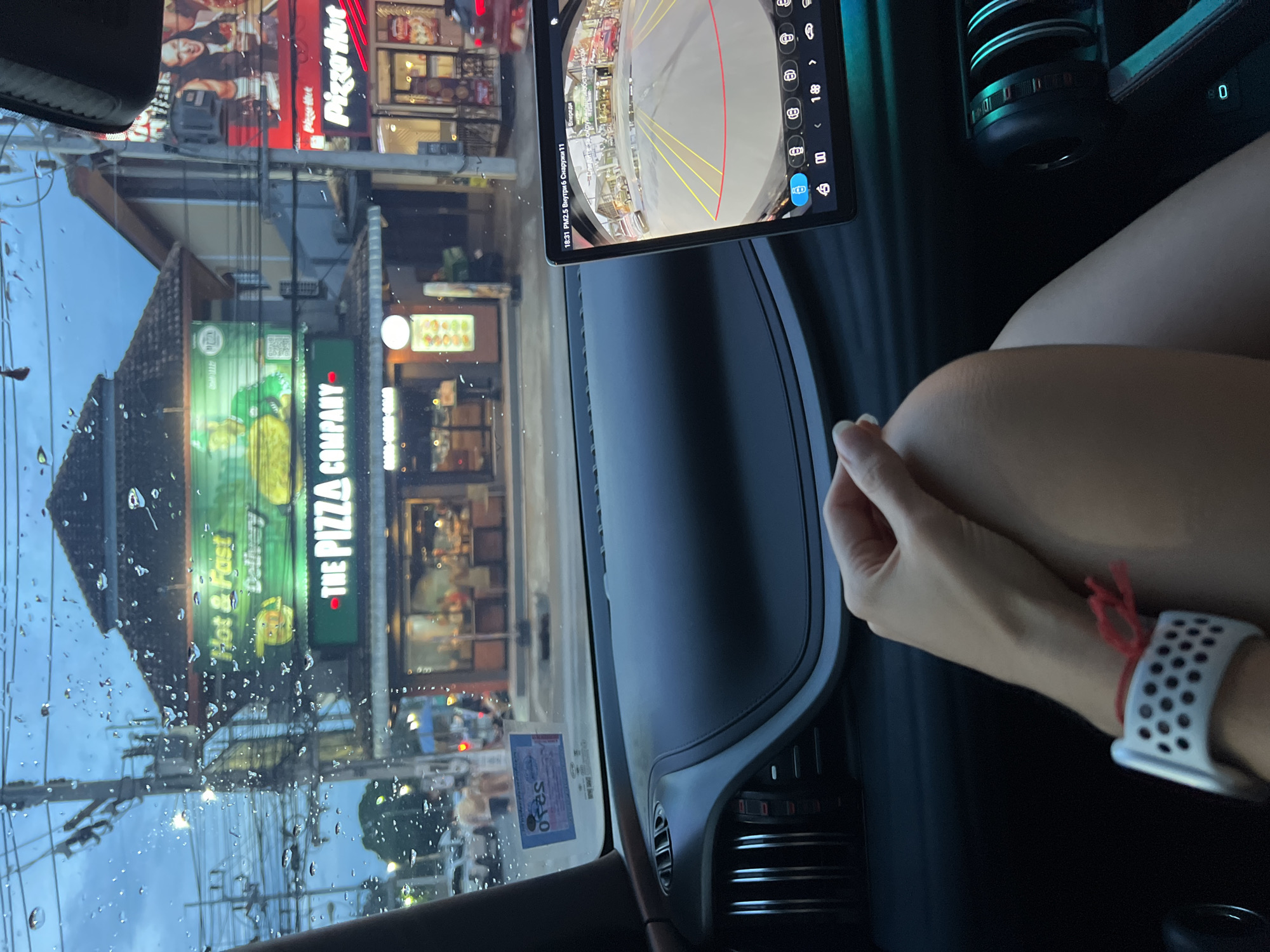 Here is a detailed description of the image:
The photograph is taken from inside a vehicle. The foreground focuses on the car's dashboard, and the view through the windshield reveals an urban street scene. Raindrops are visible on the windshield. On the left, there is a restaurant sign that reads "THE PIZZA COMPANY". Adjacent to it, a Pizza Hut restaurant can be seen.
A tablet is mounted on the dashboard displaying a rear camera view, suggesting that the vehicle is backing up. To the right, a fair-skinned person's arm and part of their leg are visible. They are wearing a white sports watch with a red band tied around the wrist.
The car's dashboard is dark, with vents and other interior elements visible. The tablet display indicates "18:31 PM2.5" alongside environmental data, and a speed of '0'.
