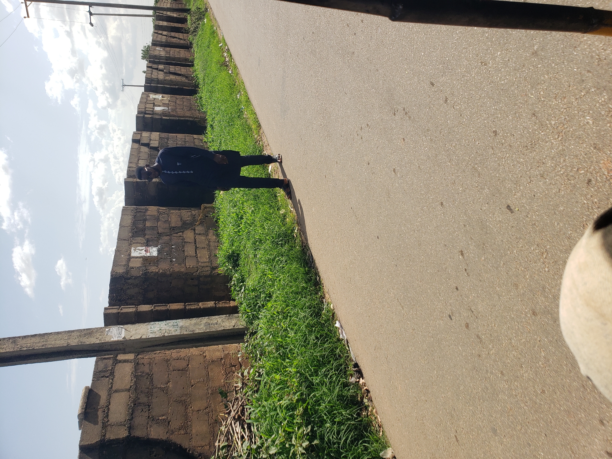 A man in a dark blue traditional outfit stands on the side of a paved road in Gura Top, Nigeria. The man is facing away from the camera, looking towards a brick wall that runs along the left side of the road. The wall is partially covered in green grass and weeds. The sky is cloudy, and there are power lines visible in the background. A white object, possibly part of a vehicle, is in the foreground. The man is wearing sandals.