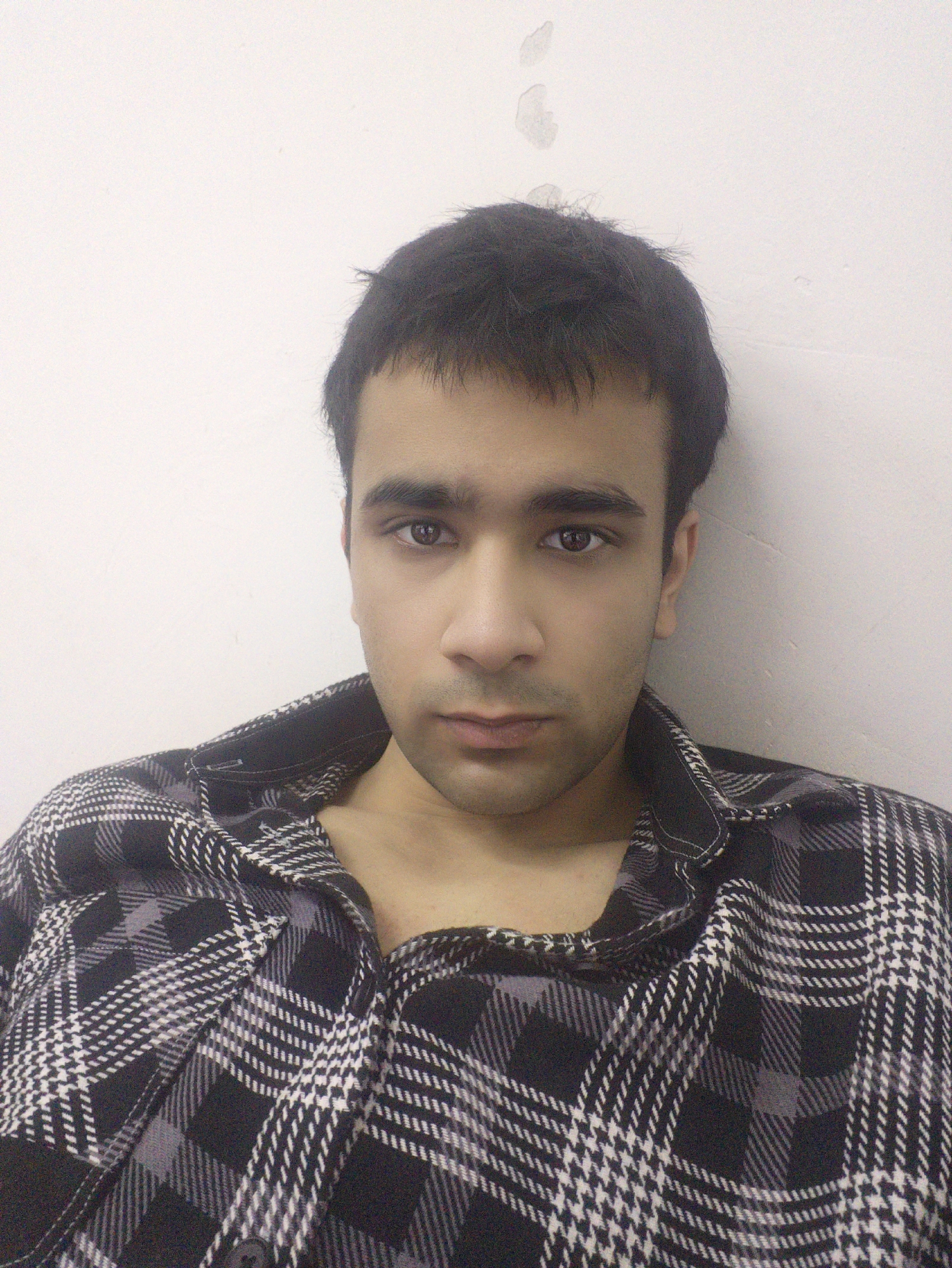 A close-up portrait of a young man with dark hair and eyes, wearing a black and white plaid shirt. The background is a plain white wall with some minor discoloration or marks in the upper portion. No evidence to suggest that the photo was taken in Chongqing High-tech Zone, China. The provided information does not give a location.