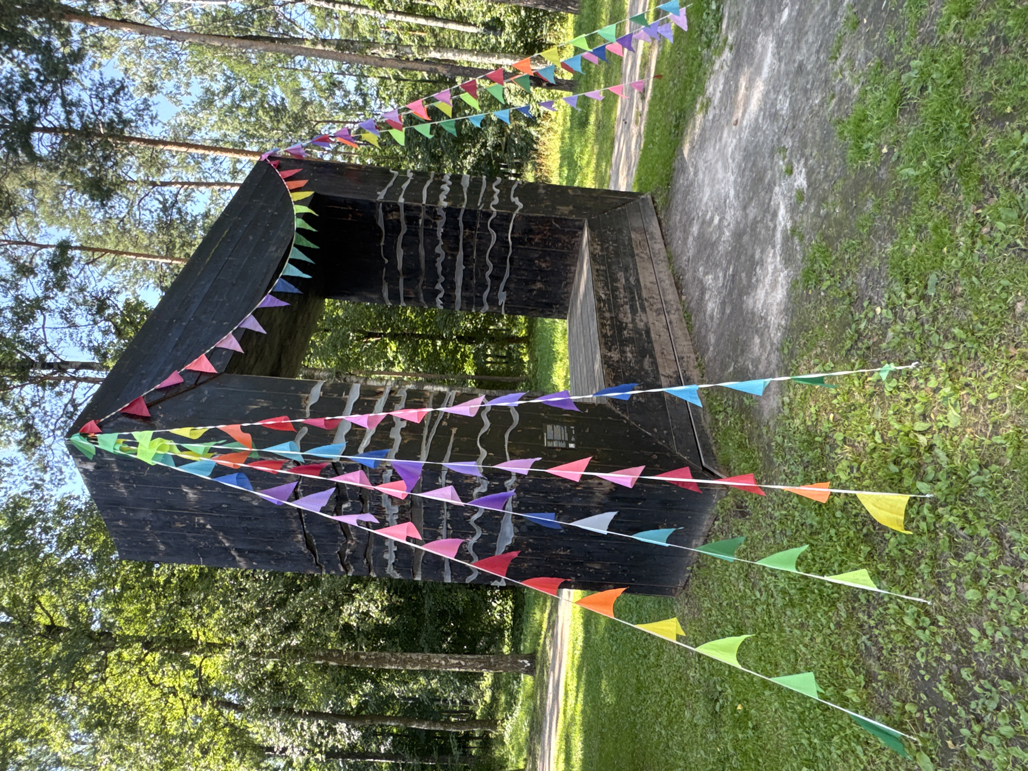 This is a full, eye-level shot of an outdoor art installation in a park setting, likely within the city of Saint Petersburg, Russia. The installation features a large, dark, angular structure made of wood, with a textured surface. Several strings of colorful triangular flags are draped across and around the structure, adding a festive atmosphere. The flags are arranged in varying patterns, some hanging straight down, others stretched taut. The surrounding environment is a natural park with lush green grass, mature trees with dense foliage, and a paved pathway. The lighting suggests it is daytime, with sunlight filtering through the trees. There are no people visible in the image. The composition focuses on the art piece and its integration with the natural surroundings.