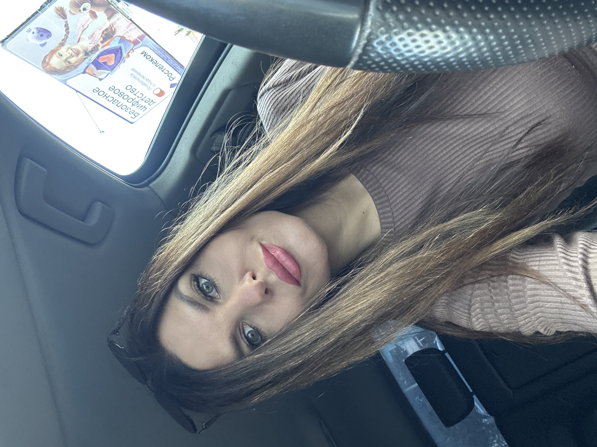 A medium shot captures a woman with long, brown hair and a mauve, ribbed sweater in the driver's seat of a car. She is looking directly at the camera with bright green eyes and pink lipstick. Her sunglasses are perched on her head. A sticker with Russian text and an image of a young girl holding a teddy bear is affixed to the car's sunroof. The sticker advertises "Safe Digital Childhood" from Rostelecom. The background is blurred, showing the interior of the car and a glimpse of the sky. The image appears to have been taken outdoors in daylight. The provided metadata indicates the image was taken in Chaikovsky, Russia.