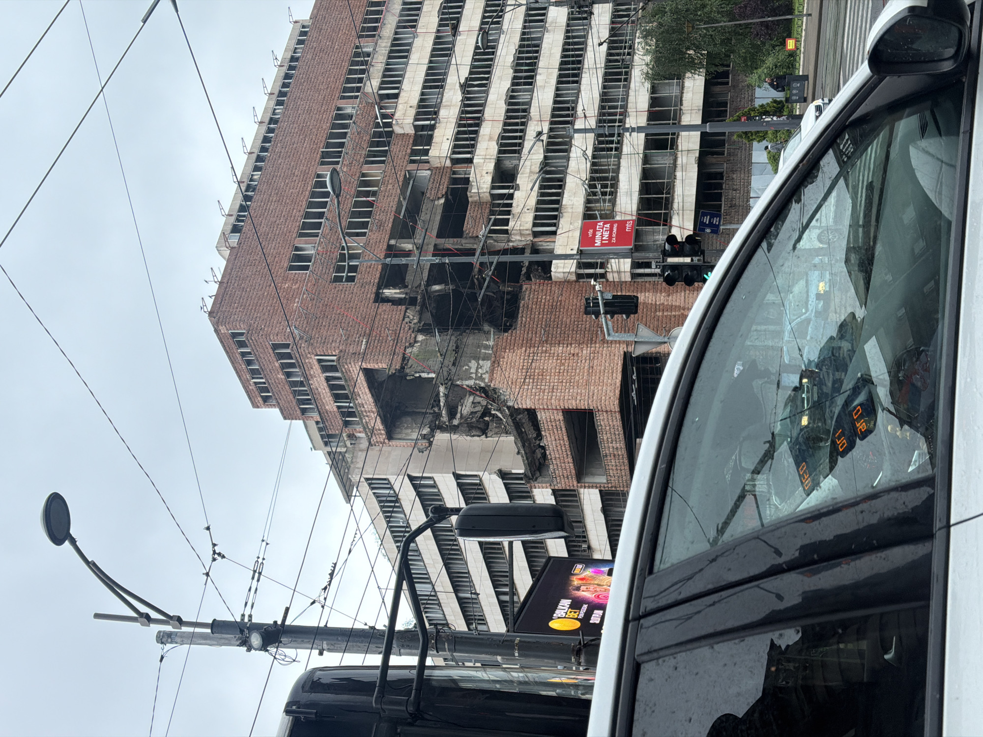 The image shows a damaged building in Belgrade, Serbia. A significant portion of the building's facade, located at its mid-section, has collapsed, leaving exposed floors and debris. The building itself has a repetitive pattern of horizontal white bands alternating with rows of windows set into a red brick facade. In the foreground is the side of a vehicle. Traffic lights are visible in the middle ground, along with signage. The sky appears overcast.