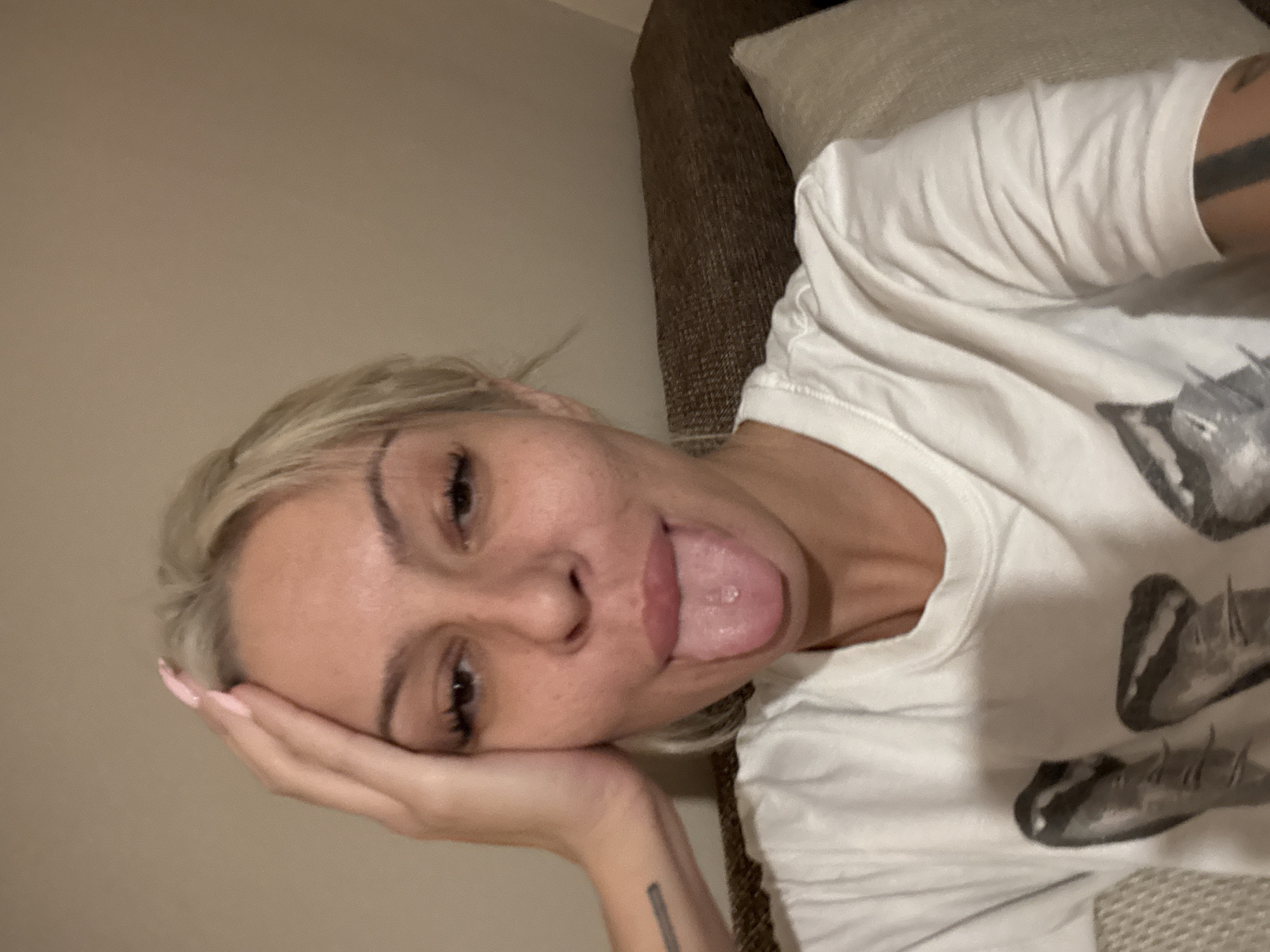A blonde woman with fair skin and visible tattoos on her arm and wrist lies on her side, partially supported by a brown upholstered couch and a light-colored pillow. She wears a white t-shirt with a printed design. Her face is close to the camera, and she has her hand supporting her head. Her eyes are dark, and she is sticking her tongue out. The background consists of a neutral-colored wall and the couch. The image doesn't give any indication that it was taken in Plovdiv, Bulgaria.