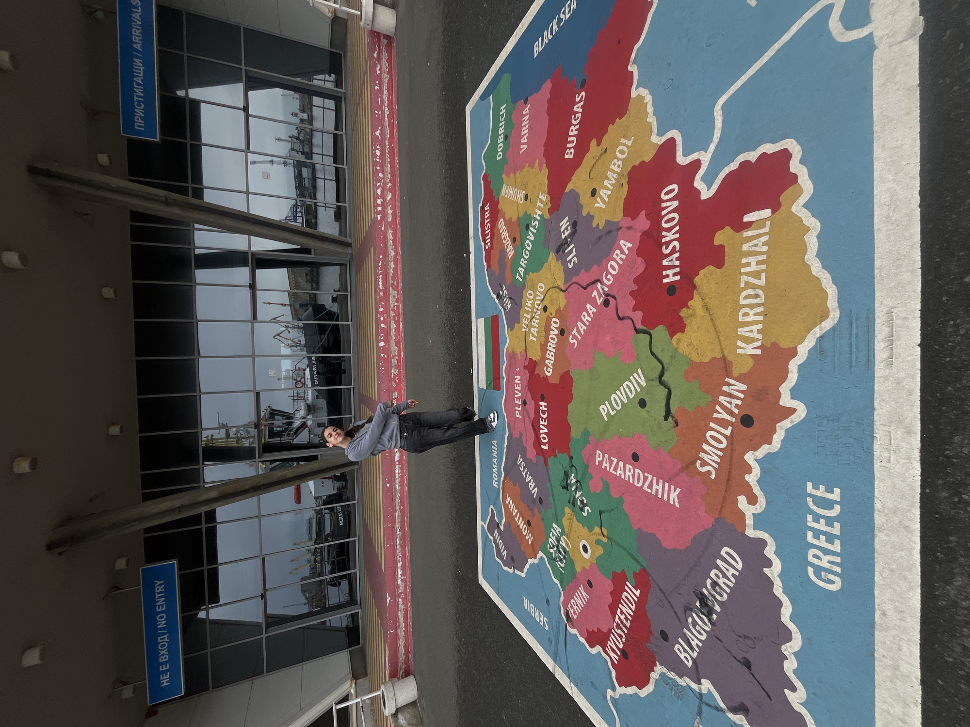 The image shows a young woman standing on a large, painted map of Bulgaria with the Black Sea and Greece also labeled. The map is located on the ground outside a building with glass windows. Signs in Bulgarian and English indicate the presence of an arrivals area. The map details the different regions of Bulgaria, including cities such as Burgas, Varna, Plovdiv, and Sofia. The environment appears to be outdoors, possibly a port area in Burgas, Bulgaria. The woman is posing for the photograph.