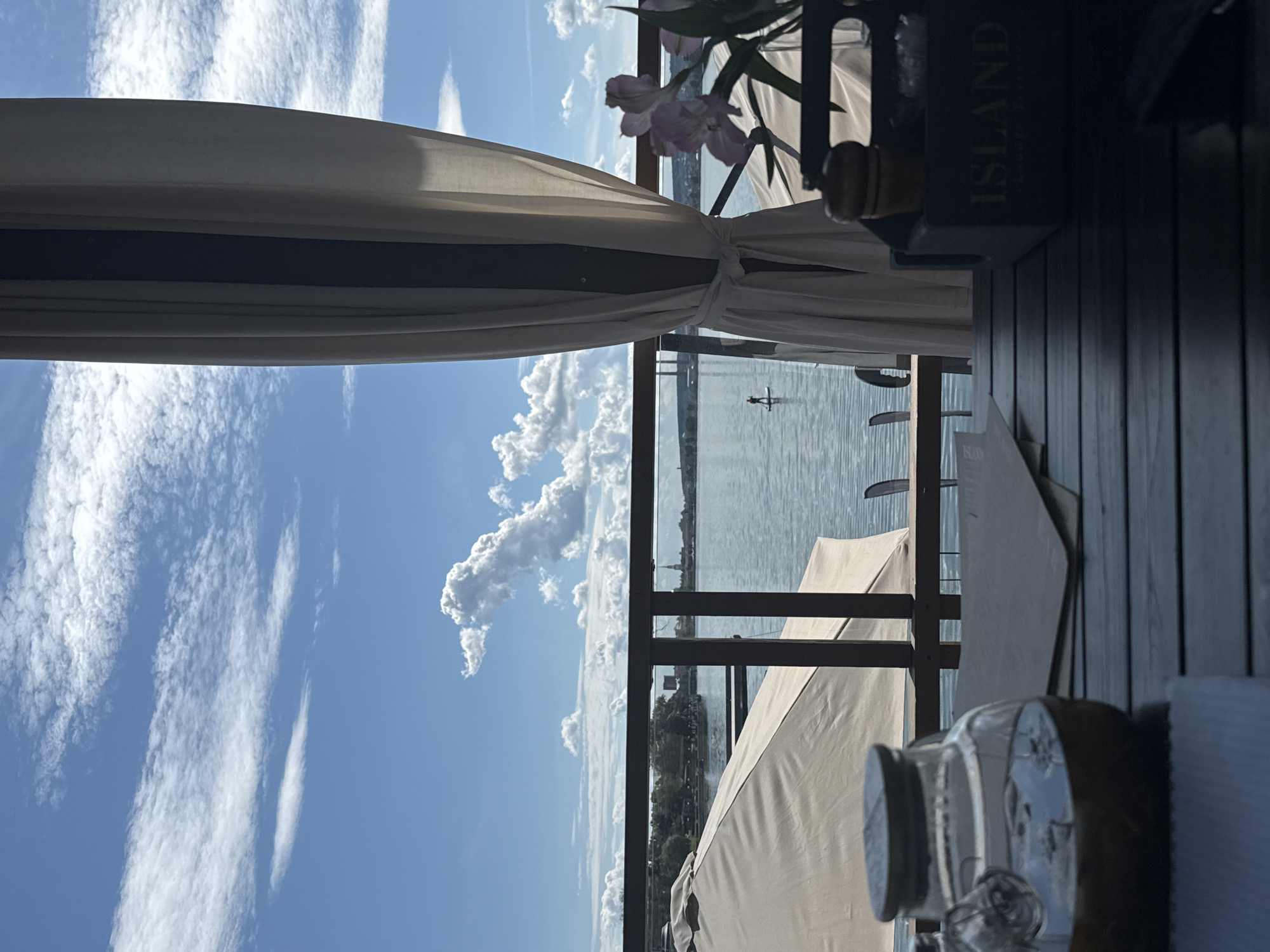 A horizontal, medium shot captures an outdoor seating area at a waterfront establishment, possibly a restaurant or cafe, overlooking a body of water under a bright blue sky filled with scattered white clouds. In the foreground, a dark wooden table is visible, with a glass carafe containing an amber liquid on the left side. To the right, a grey caddy holds salt and pepper shakers and a small container with white contents. Partially obscured by a white curtain in the center right, a vase with delicate pink flowers is positioned on the table.
The middle ground reveals a railing separating the seating area from the water. Beyond the railing, several large, beige umbrellas are set up, suggesting outdoor dining or lounging spaces. The water is calm, reflecting the sky and distant shoreline. A lone figure is seen paddleboarding on the water, adding a touch of activity to the serene scene.
The background shows a cityscape with buildings and infrastructure, including a bridge in the distance. The location is identified as Votkinsk, Russia. The overall atmosphere is tranquil and leisurely, with a focus on the connection between the seating area and the natural environment.