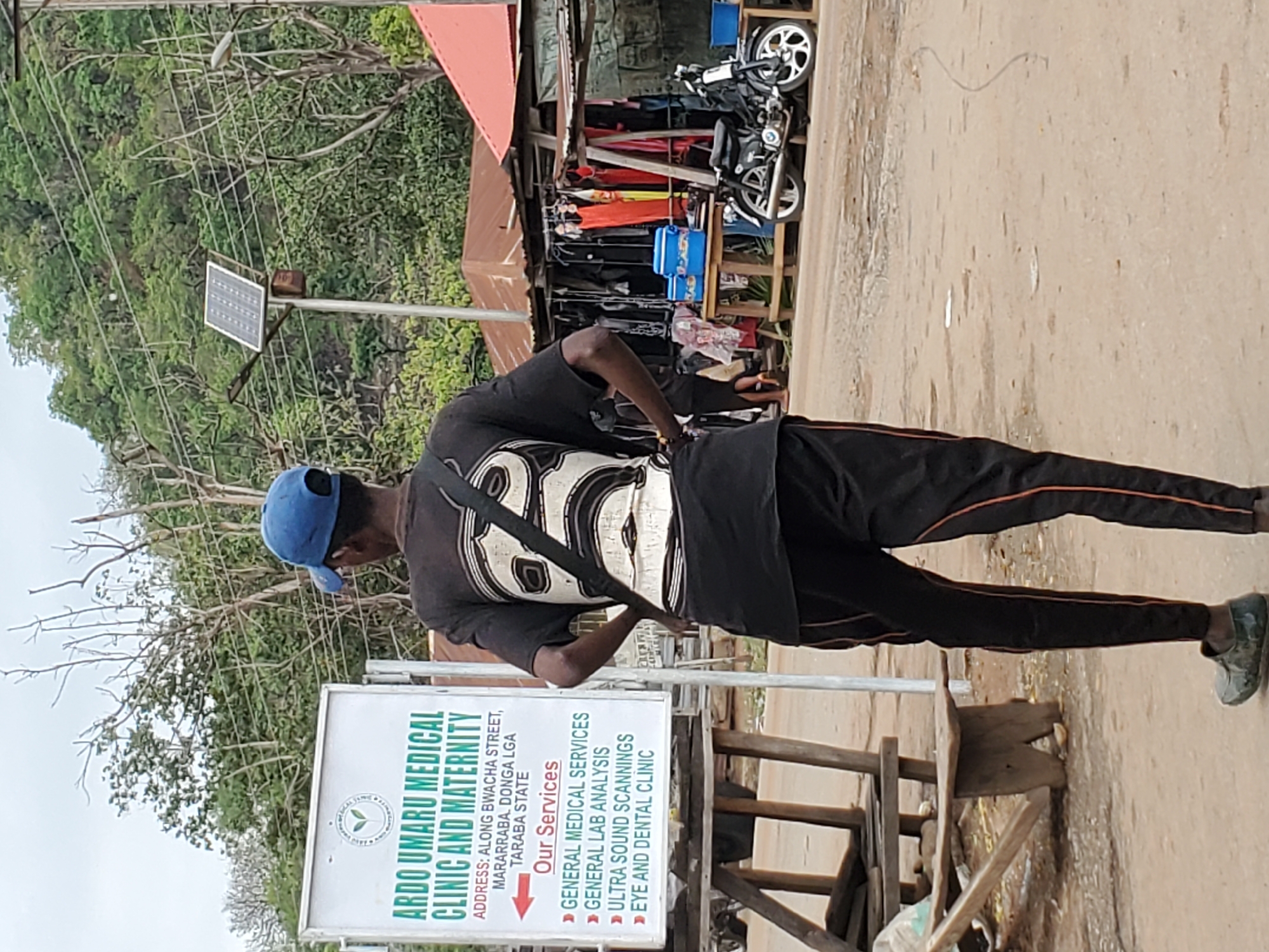 The image captures an outdoor street scene, likely in Mararaba, Donga LGA, Taraba State, Nigeria, as confirmed by text on a visible sign. In the foreground, a man is seen from behind, standing next to a white medical clinic sign. He wears a blue baseball cap, a black t-shirt with a large white graphic (resembling "86"), black track pants with orange side stripes, and dark shoes, with a black bag slung across his body.
The sign, prominently displayed, reads "ARDO UMARU MEDICAL CLINIC AND MATERNITY" and provides the address: "ALONG BWACHA STREET, MARARRABA. DONGA LGA TARABA STATE." Under the heading "Our Services," it lists: "GENERAL MEDICAL SERVICES," "GENERAL LAB ANALYSIS," "ULTRA SOUND SCANNINGS," and "EYE AND DENTAL CLINIC."
In the background, to the left, a lush green hillside or dense trees are visible, along with a utility pole featuring a solar panel and a street light. Several overhead power lines crisscross the upper portion of the image. To the right of the man, across an unpaved road, is a rustic roadside stall or shop, with clothes hanging for sale, some blue containers, and a black motorcycle parked. The ground is a light brown, possibly dirt or gravel. The scene is illuminated by daylight under what appears to be a clear or lightly overcast sky.