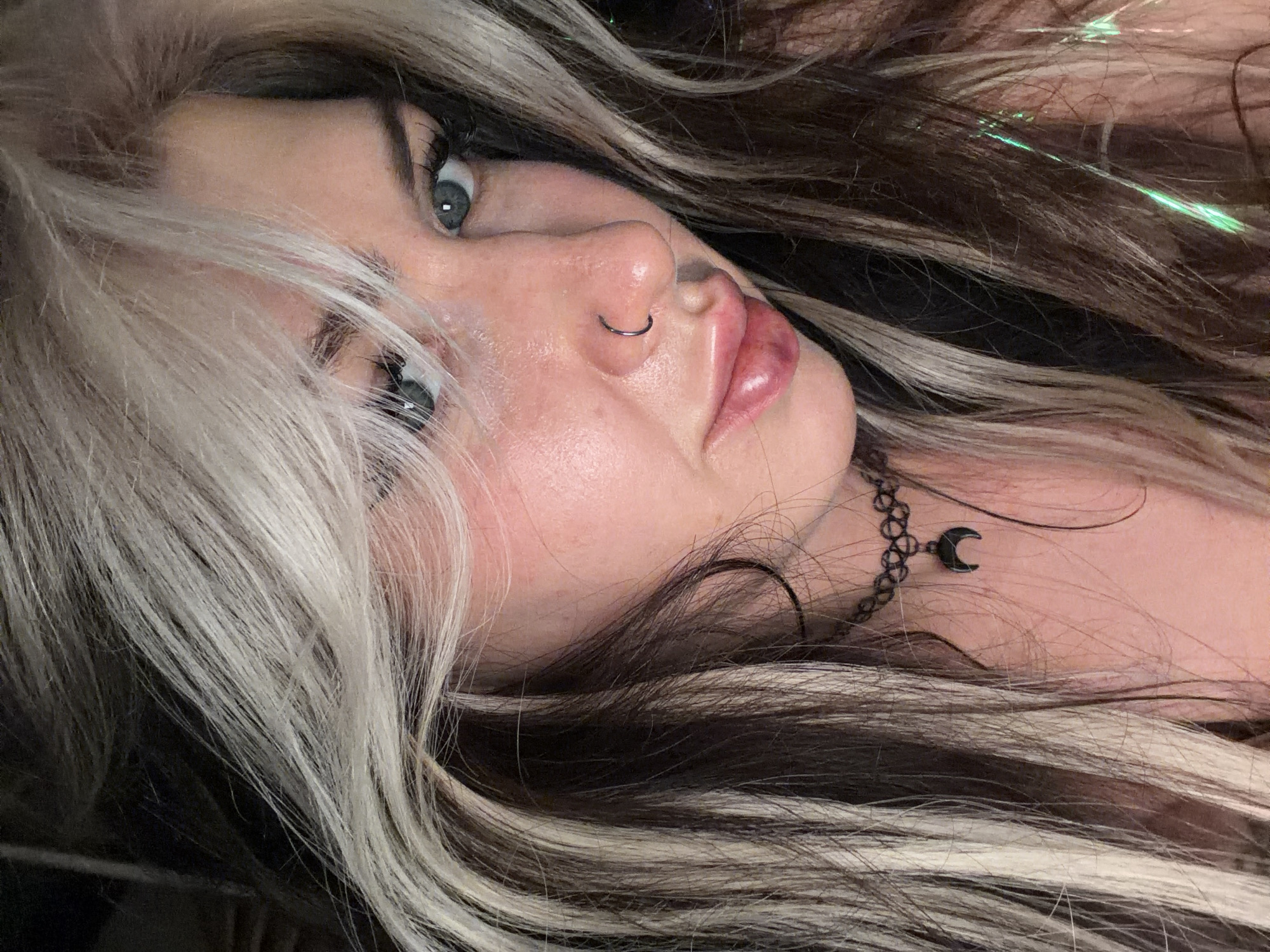 A close-up, angled portrait shows a woman with dyed black and white hair. She has fair skin, blue eyes, and a nose ring. She wears a black choker with a crescent moon pendant. A thin, green, sparkling strand is woven into her hair. The background is out of focus and dark. The city location in Budva, Montenegro is not evident in the provided image.