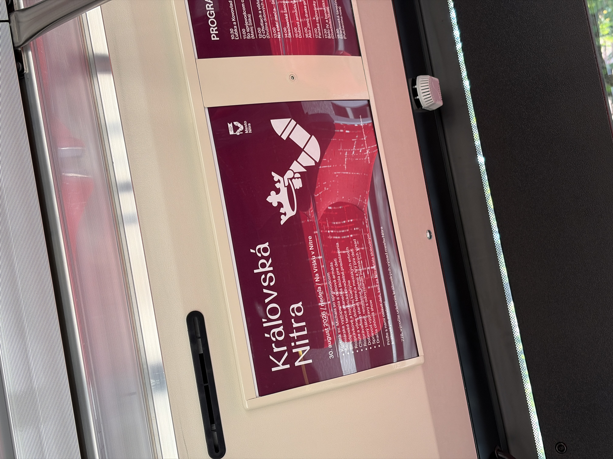 The image shows a close-up of a digital display screen mounted inside a public transport vehicle, likely a bus or tram, with the overhead interior visible. The display advertises an event called "Kráľovská Nitra" (Royal Nitra) taking place on August 30, 2020, on "Na Vŕšku" (on the hill/top) in Nitra. The program lists various activities such as a guided tour, dance performances, historical reenactments, and a show. The city emblem of Nitra is visible on the display, featuring a crown. The display is illuminated by the ambient light within the vehicle and appears to be a promotional advertisement.