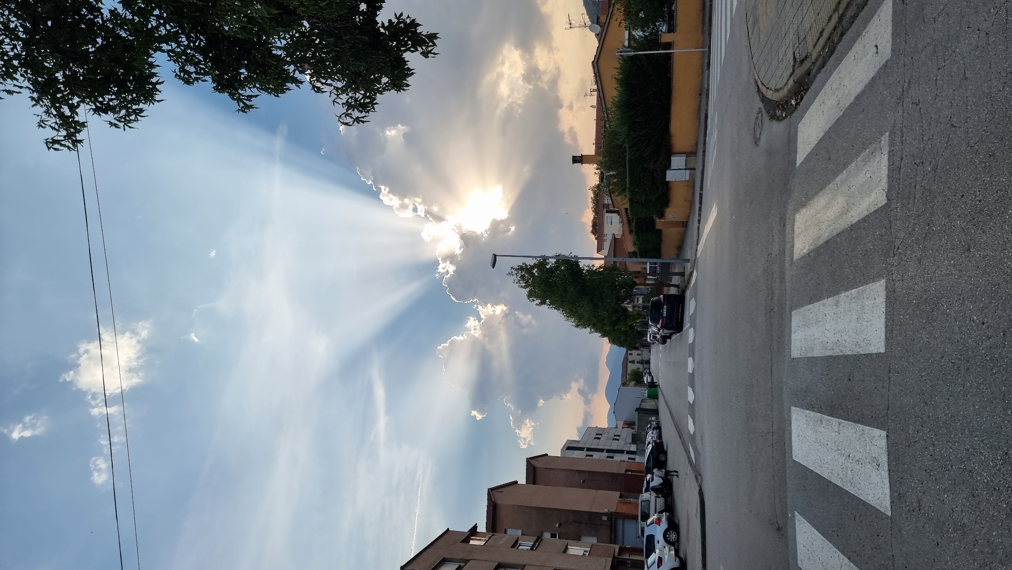 The image captures a street scene in Sant Joan Les Fonts, Spain, featuring a pedestrian crossing in the foreground and a street lined with parked cars and buildings. In the background, a dramatic sky is illuminated by crepuscular rays emanating from behind a large cloud formation. The sun is setting, casting a warm glow over the scene. On the left, multi-story residential buildings are visible, while on the right, a building with an orange facade and manicured hedges lines the street. Trees are planted along the sidewalk. Several vehicles are parked along the street on both sides. The sky is a mix of blue with scattered white clouds, highlighted by the sun's rays.