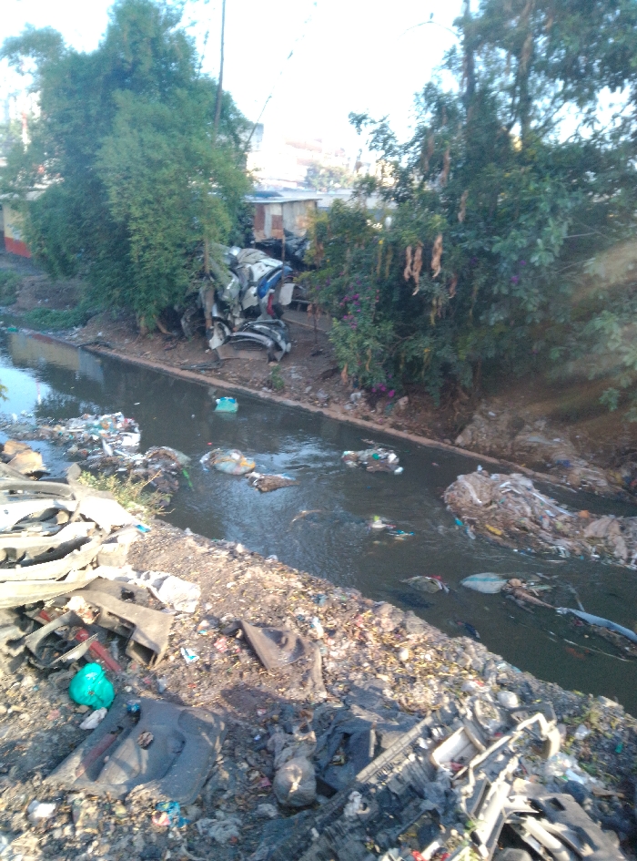 The image portrays a heavily polluted waterway, likely a river or canal, within an informal settlement in Nairobi, Kenya. The water is dark and appears turbid, carrying numerous pieces of floating refuse including plastic containers, bags, and unidentifiable debris. Both banks of the waterway are extensively covered with accumulated garbage. In the foreground on the left bank, a large mound of waste is visible, consisting prominently of discarded automotive components such as bumper covers and interior panels, intermingled with plastic bags and general household refuse. On the opposing bank, similar clusters of trash are present, including a substantial pile of light-colored debris near the water's edge and a stack of automotive body parts further back, situated near several rudimentary structures. These structures are partially obscured by dense green foliage and appear to be makeshift dwellings constructed from corrugated metal and other salvaged materials. Various trees and shrubs, some exhibiting long, hanging seed pods, line the banks. No individuals are visible in the scene. The sky is bright overhead, suggesting daylight conditions.