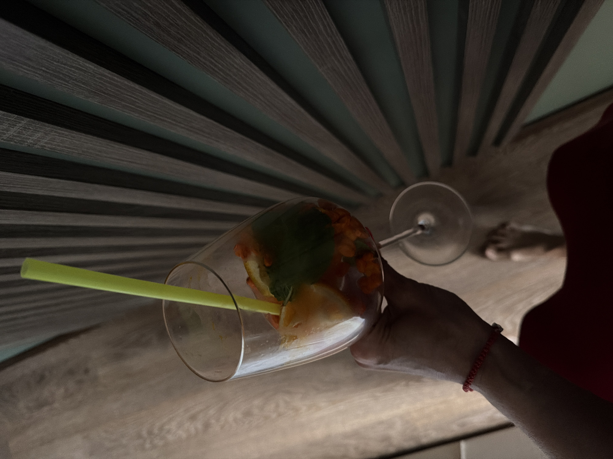 In Birobidzhan, Russia, a first-person perspective photograph captures a bare left forearm and hand holding a stemmed wine glass. The wrist is adorned with a red braided string bracelet. Inside the glass, a clear liquid contains small, round, orange berries (resembling sea buckthorn), a citrus fruit slice (possibly orange or lemon), and several green mint leaves. A bright yellow plastic straw protrudes from the beverage. The immediate foreground shows a light brown wooden floor. The background features a distinctive wall panel composed of alternating vertical strips of dark wood-grain material and a muted teal-green solid color, arranged in a radial pattern that appears to converge towards the bottom-center of the panel. A blurred bare foot and a portion of a dark red garment are visible in the lower left of the frame.