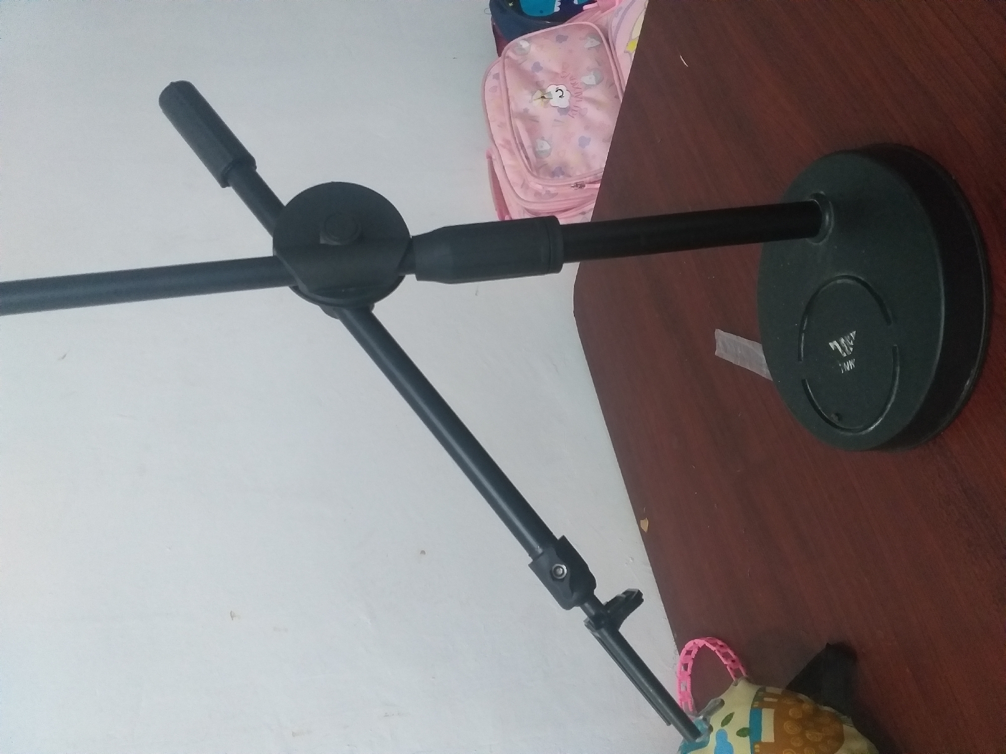A black microphone stand with a round base is positioned on a wooden table. The stand has an adjustable boom arm that extends diagonally to the left. In the background, a white wall is visible, with a pink backpack adorned with cartoon unicorns and a blue backpack partially in view. A plush toy with a brown and yellow pattern and a pink plastic ring is also on the table to the left of the stand. The scene appears to be indoors.