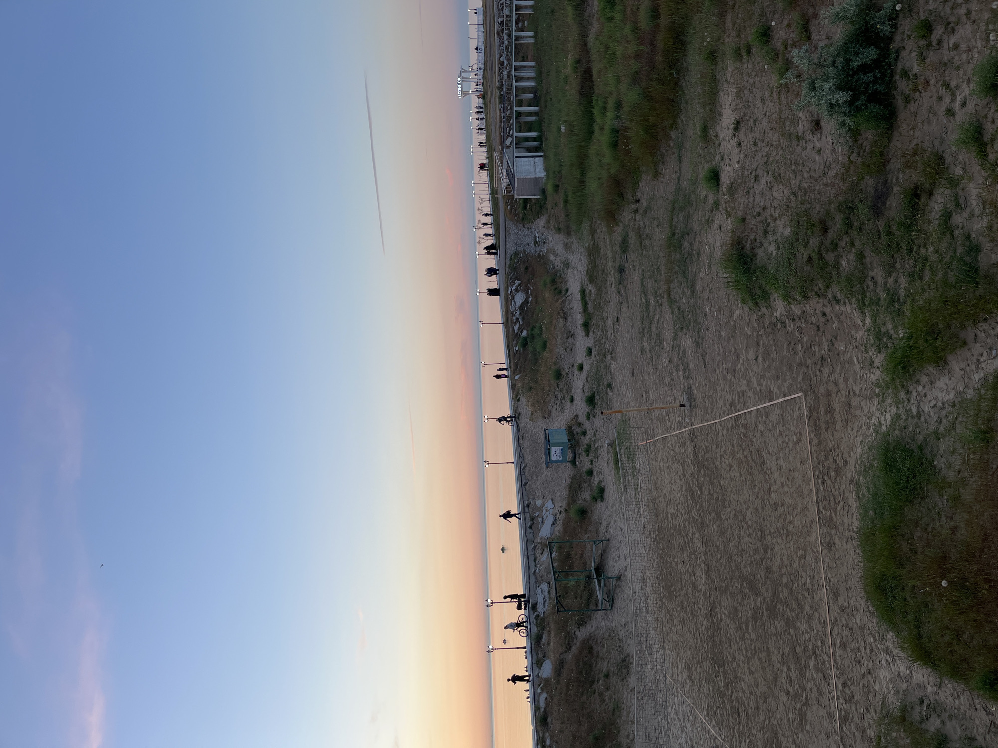 The image shows a view of the beach in Kaspiysk, Russia, at dusk. The sky is a gradient from blue to orange near the horizon. A contrail is visible in the sky. Along the waterfront, there are lampposts and people walking and biking. A path runs along the shore, bordered by grass and vegetation. On the beach, a volleyball net is set up.