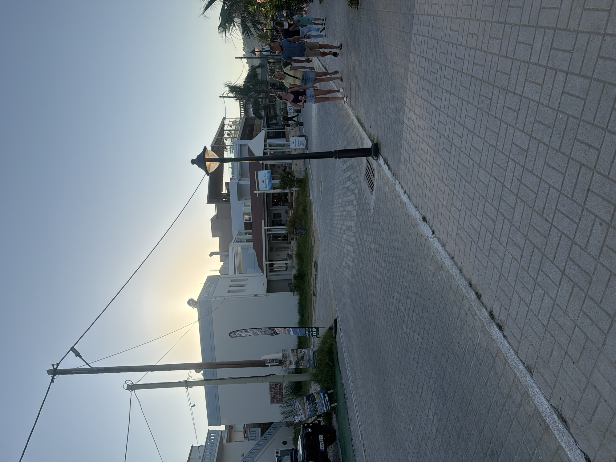 The image shows a street scene in Georgioupoli, Greece, during daylight hours with the sun low in the sky. A paved street runs diagonally from the bottom left to the upper right of the frame, with a sidewalk on the right side. Several buildings line the street, some with shops or restaurants on the ground floor. A dark-colored SUV is parked on the left side of the street. Two utility poles with wires extend across the sky. Several people are walking on the sidewalk. The overall scene depicts a quiet street with a few pedestrians and vehicles, suggesting a relaxed atmosphere.