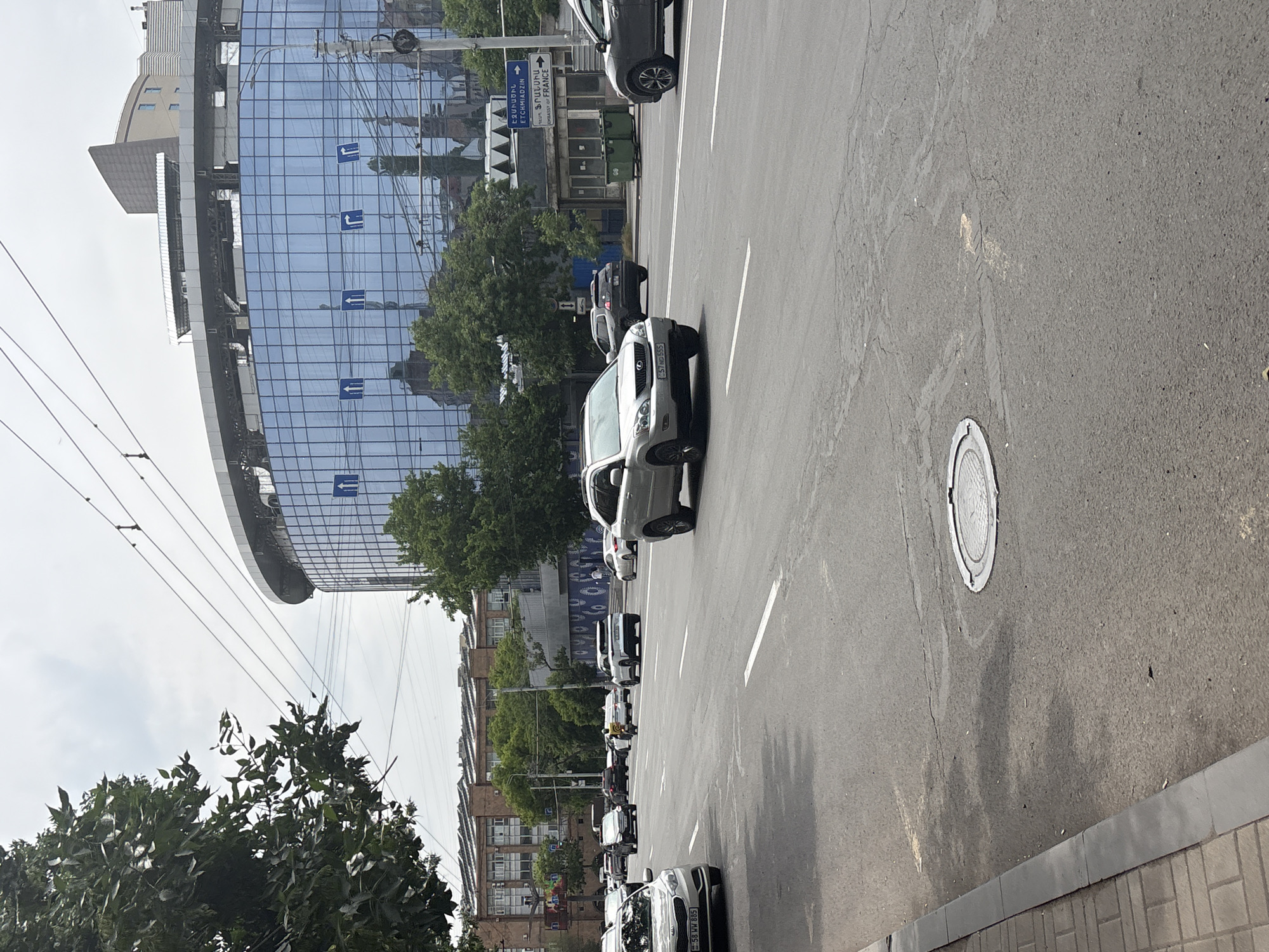 The image captures a street scene in Yerevan, Armenia. A silver SUV is centered in the view driving down the street. Numerous other cars line the side of the street. The road itself is marked with white lane dividers and a circular manhole cover. A modern, curved glass building with arrow symbols on the facade and a street sign indicating directions, including "Embassy of France", are also visible. Trees line the streets. The weather appears overcast.