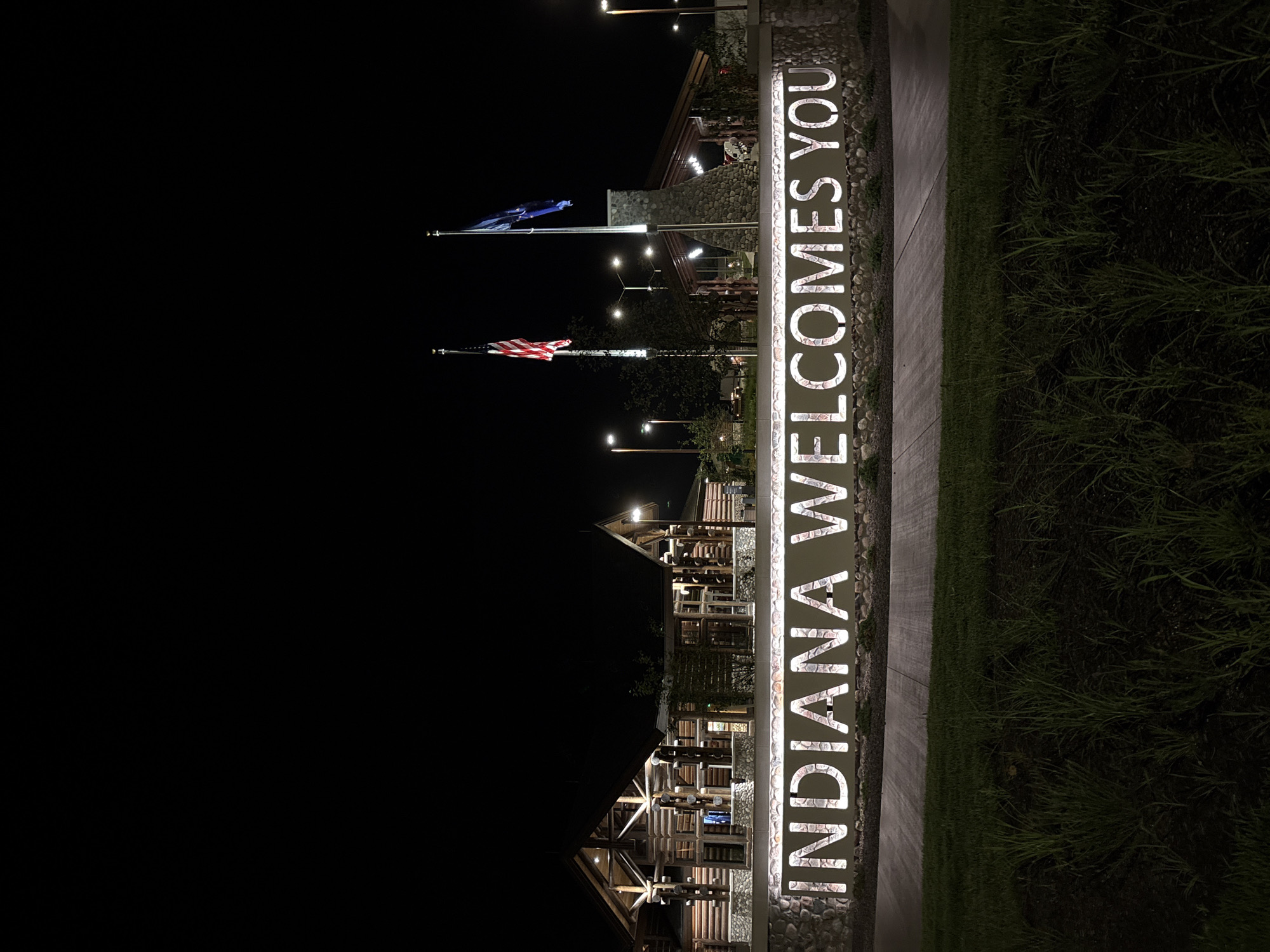 This image captures the Indiana Welcome Center at night, located near New Harmony, Indiana, United States. The scene is illuminated by artificial lights, highlighting a large, illuminated sign that reads "INDIANA WELCOMES YOU." The sign is mounted on a stone wall and is backed by a textured pattern.
Behind the sign, the Welcome Center buildings are visible, constructed with log cabins and stone elements, suggesting a rustic architectural style. Two flagpoles stand prominently in front of the buildings, one displaying the American flag and the other a blue flag, possibly representing Indiana or another state entity. Several lampposts are also visible, providing additional lighting to the area. In the foreground, there is a patch of grass with some plants. The overall impression is one of a welcoming gateway to the state, observed at night. There are no people or animals visible in the frame.