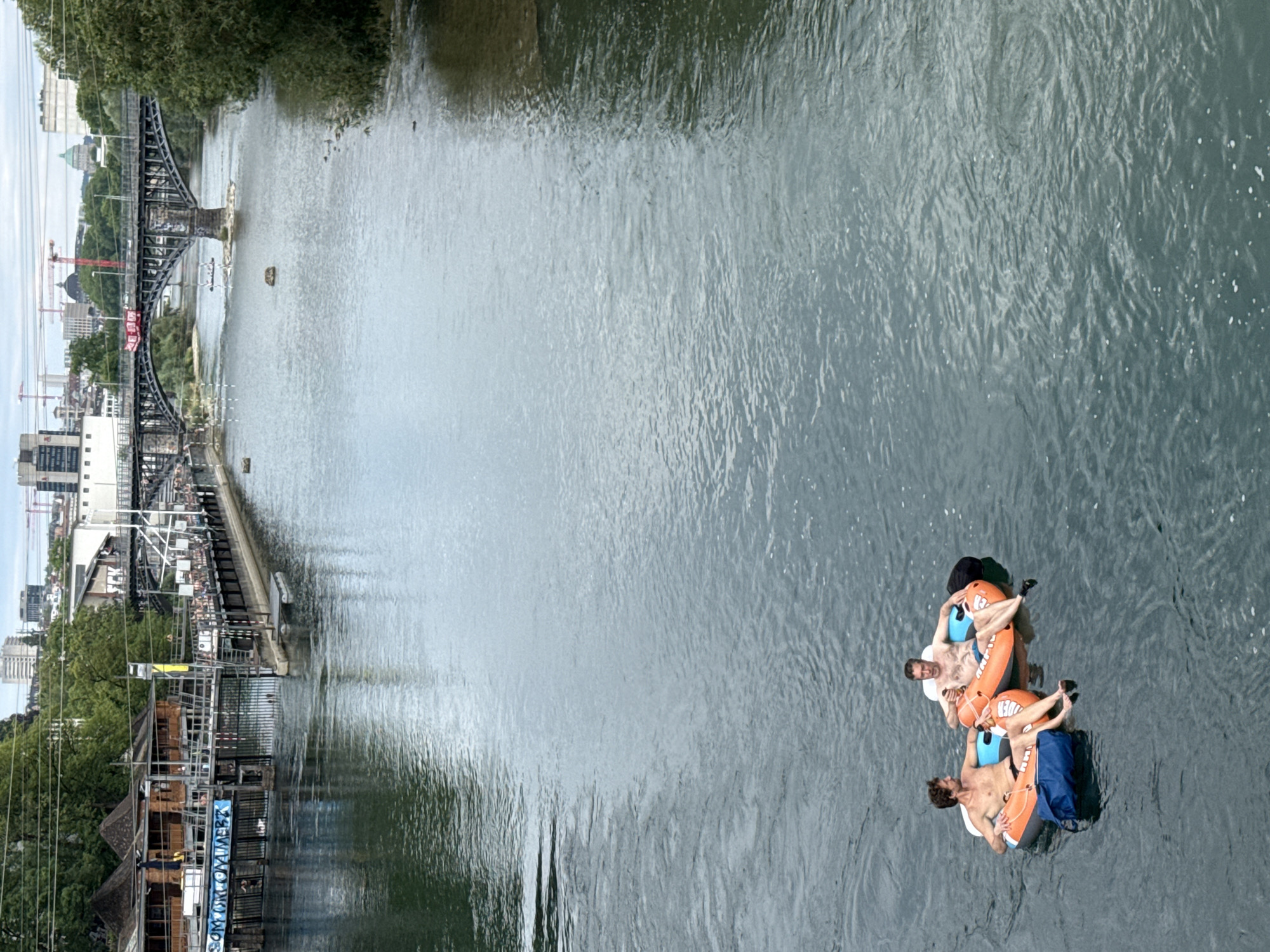 Two shirtless men are floating on inflatable tubes down the Limmat river in Zurich, Switzerland. They are in the foreground of the image, with the man on the left in an orange and blue tube and the man on the right in an orange tube. Both men are wearing swim trunks. The man on the left has a blue bag next to him in the water. The man on the right is holding a drink. In the background, there is a bridge spanning the river, with buildings and trees on either side. The buildings appear to be part of the Zurich cityscape. The water is a murky green color, and there are ripples on its surface, indicating a gentle current. The sky is overcast.
