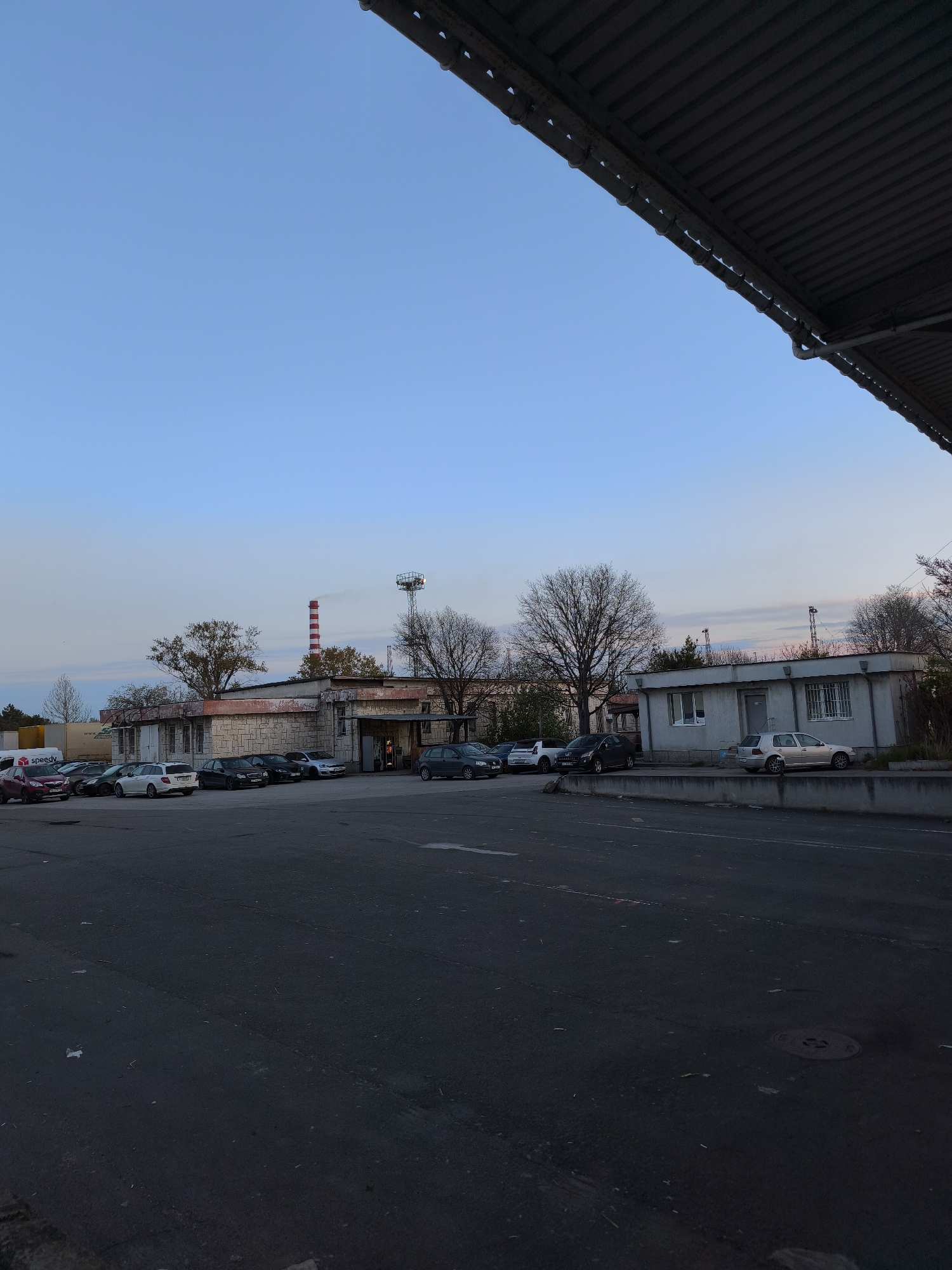 The image depicts an outdoor parking area in Burgas, Bulgaria, at dusk or early evening.
**Environment:** The sky is light blue, transitioning to a lighter shade near the horizon, with possible hints of pink/orange hues indicating sunset or sunrise. The ground is asphalt, dark in color, and appears damp in places. There's a large industrial-looking building complex in the background, consisting of various structures of different sizes and architectural styles. A tall red-and-white striped chimney is visible amongst these buildings. The angle is taken from beneath a corrugated metal overhang, casting shade in the upper right.
**Objects:** Several parked cars are scattered across the parking area. A concrete or stone barrier separates the parking area from the closest building. A manhole cover is visible in the foreground asphalt.
**People:** There is no clear indication of people in the scene.
**Location:** Burgas, Bulgaria.
**Actions:** The image is a static scene with no observable actions or interactions.