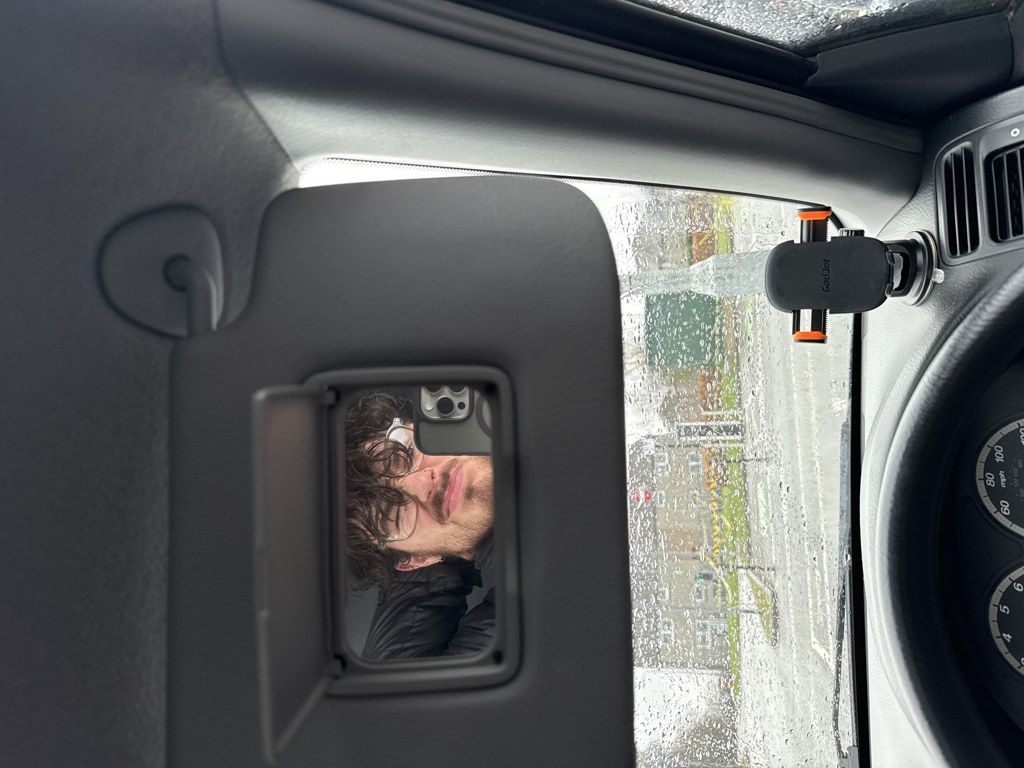 The image depicts the interior of a vehicle on a rainy day, with a man's reflection visible in the vanity mirror of the sun visor. The man has curly hair, wears glasses, and is wearing a black jacket. Outside the car window, droplets of rain obscure the view of a street with buildings and possibly a crosswalk with yellow markings. A phone mount is attached to the dashboard. The speedometer is visible, displaying a range of numbers. The location seems to be an urban area.