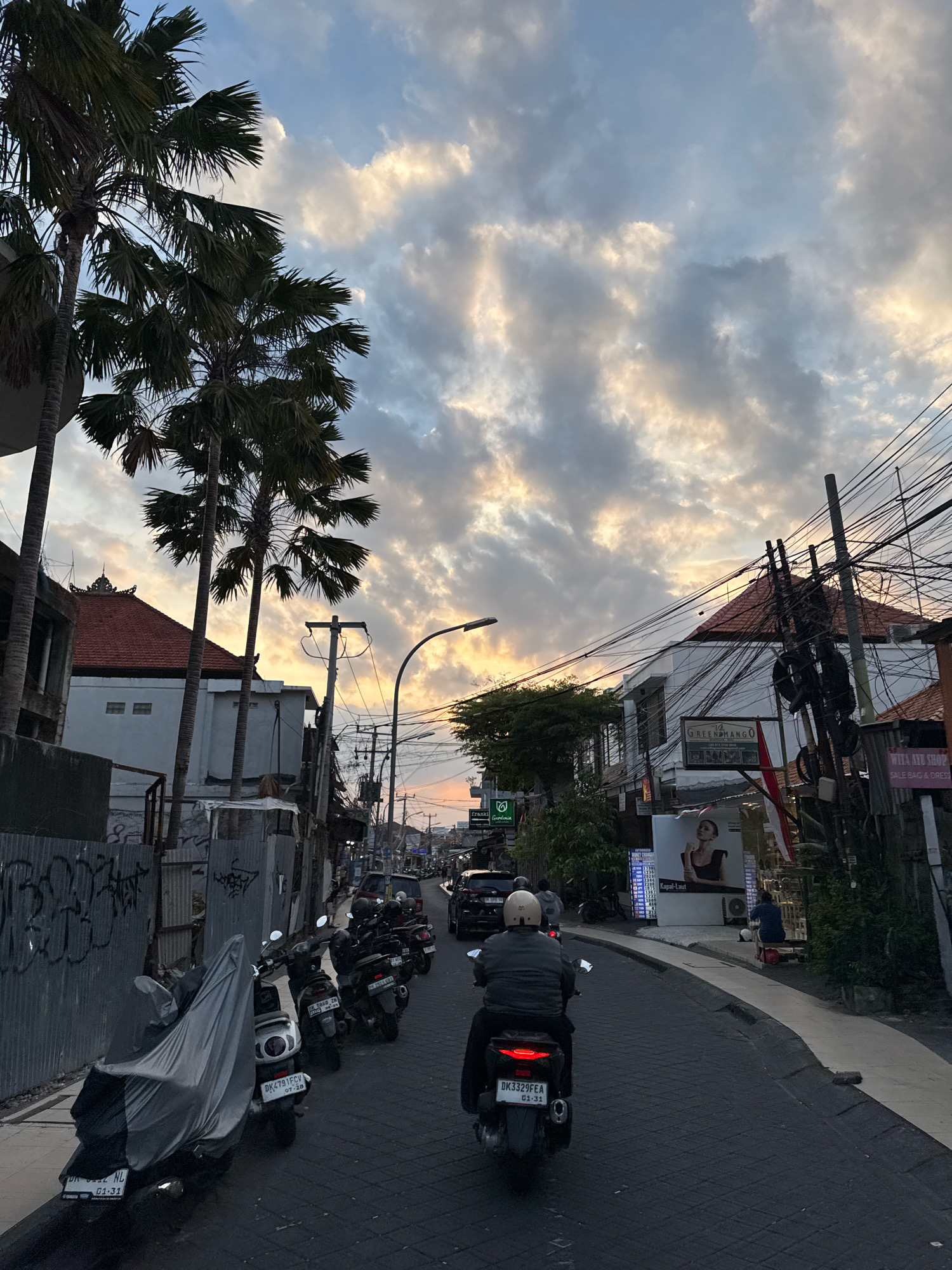 The image depicts a street scene in Kuta, Indonesia, during what appears to be either sunrise or sunset, as indicated by the warm, cloudy sky. The street is paved with dark gray stones and has a sidewalk on the right. A person is riding a black scooter with a white helmet, heading away from the viewer. The scooter has a license plate that reads "DK3329FEA 01-31". Along the left side of the street, there are several parked scooters, one of which is covered with a light gray tarp. Behind the scooters, there's a corrugated metal fence with graffiti. Buildings line both sides of the street; on the left, there's a building with a tiled, reddish-brown roof and a white facade. On the right, buildings have white walls and reddish-brown tiled roofs, with numerous electrical wires strung between poles. One building features a sign for "GREEN MANGO" and another poster with an image of a woman. Palm trees with large fronds are visible on the left side of the frame, reaching towards the sky. The overall atmosphere is that of a bustling urban environment with a tropical touch, common in Kuta.