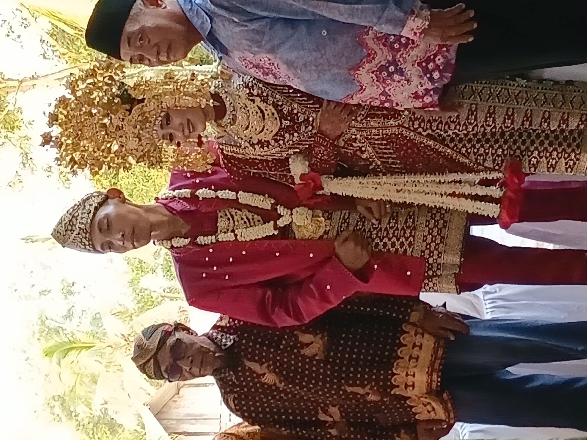 In the city of Sumber Mulyo, Indonesia, a wedding ceremony is underway. Four individuals are posing for a photograph. On the far left stands an elderly man wearing sunglasses and a dark patterned shirt with gold accents. Next to him is the groom, adorned in a red suit and a traditional headdress, with a floral garland draped around his neck. To his right is the bride, elegantly dressed in a red and gold wedding gown and a tall, ornate headdress. Completing the group on the far right is an older man in a light blue shirt with a patterned collar. The background features a rustic structure and lush greenery, suggesting an outdoor or semi-outdoor setting.