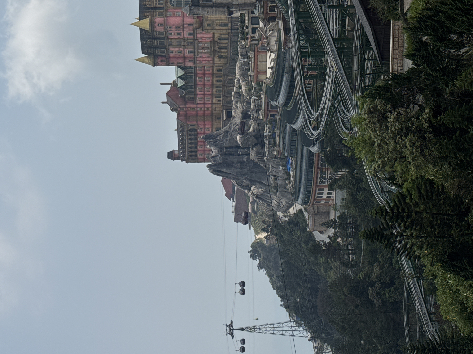 The image shows a partial view of Ba Na Hills, a mountain resort in Đà Nẵng, Vietnam. In the upper left is a pink and gold European-style building. A stone formation is visible adjacent to the building. Rollercoaster tracks and support structures run along the upper right side. Trees and green foliage are visible in the lower right. A cable car system, consisting of towers and gondolas, is visible in the lower portion of the image, descending towards the mountain base. The sky is pale gray and cloudy.