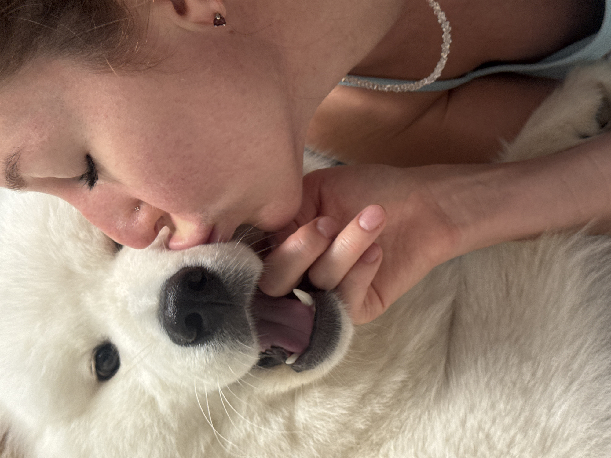 A close-up, eye-level shot shows a person kissing a white Samoyed dog on the muzzle. The person's eyes are closed, and their head is tilted to the left. They are wearing a light blue camisole and a beaded necklace. The dog's mouth is open, revealing its pink tongue and white teeth. The background is blurred, but appears to be indoors, possibly in a home. There are no identifying landmarks or elements that would place the image in Chaikovsky, Russia.