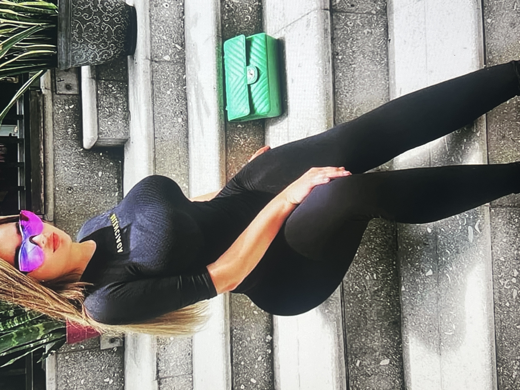 A woman with long blonde hair and large pink sunglasses is sitting on outdoor concrete steps. She is wearing a black full-body outfit and her hands are resting on her thighs. A bright green quilted purse is placed on the step next to her. In the background, a large potted plant with striped leaves is visible to the left, and another decorative pot is to the right. The scene appears to be an outdoor setting, possibly a public space with stairs. The image does not contain any information about the city of Vanadzor, Armenia.