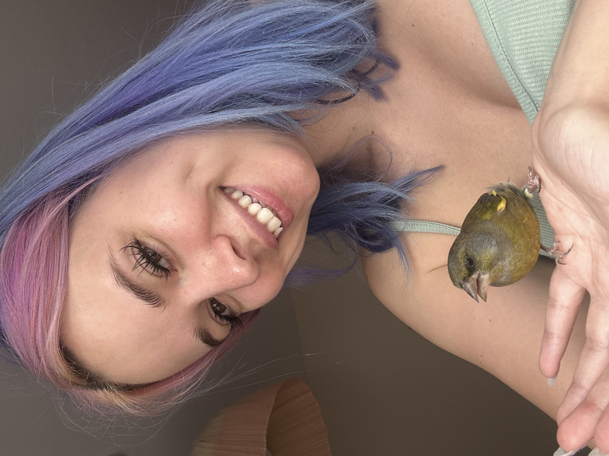 A young adult female, presenting with bi-colored pink and lavender hair, is depicted smiling directly towards the viewer. Her face shows brown eyes, prominent dark eyelashes, and a wide smile revealing upper and lower teeth. She is wearing a light mint green ribbed camisole. Her left hand is extended and open, supporting a small bird. The bird, exhibiting olive-green and yellowish plumage with a short, conical beak, is perched calmly on her palm and fingers, facing slightly left with its beak slightly agape. The setting is indoors, with a blurred, neutral light brown background, and a partial view of a curved, light orange-brown fixture in the upper left quadrant. The scene takes place in Gijón/Xixón, Spain.